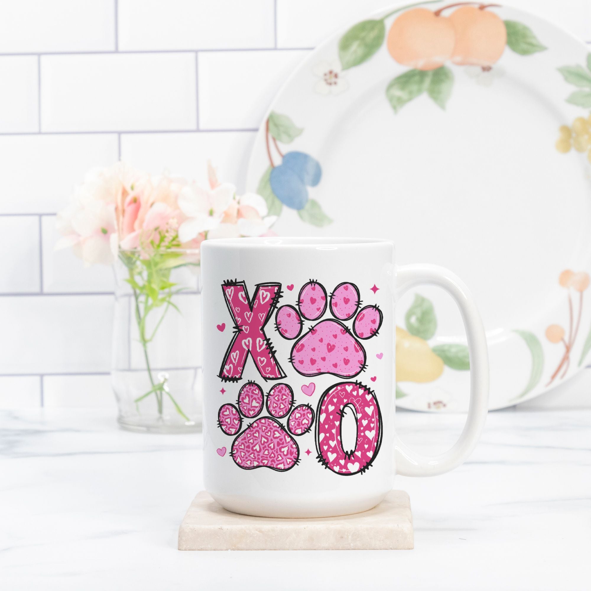 White mug with pink paw print design on a white surface with a floral plate in the background.