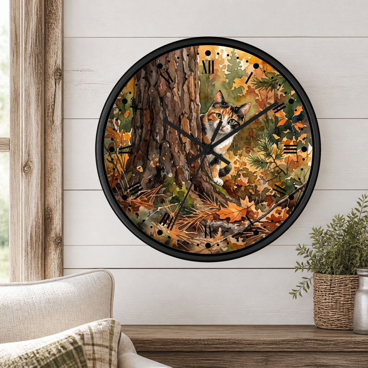 A round wall clock with a black frame and a printed watercolor autumn forest design featuring a peeking calico cat, mounted on a white shiplap wall above a wooden mantel with a plant and cushion.