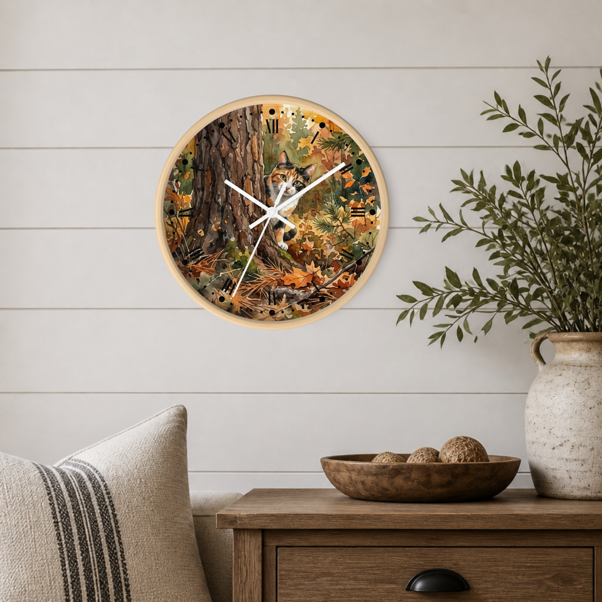 A round wall clock with a light wood frame hangs on a white shiplap wall above a wooden side table. The clock face features a detailed autumn forest scene with a peeking calico cat behind a tree among fallen leaves.