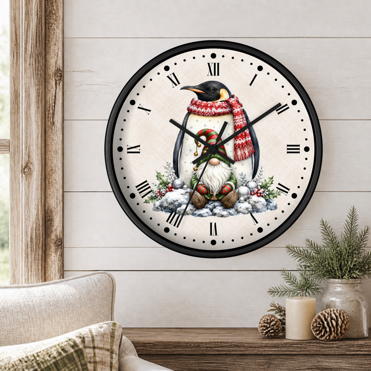 A round wall clock with a black frame and a white face, featuring a penguin illustration wearing a red and white striped scarf and a green hat, set against a snowy background. The clock is mounted on a light wood-paneled wall above a wooden table with a candle, pinecones, and a small plant.