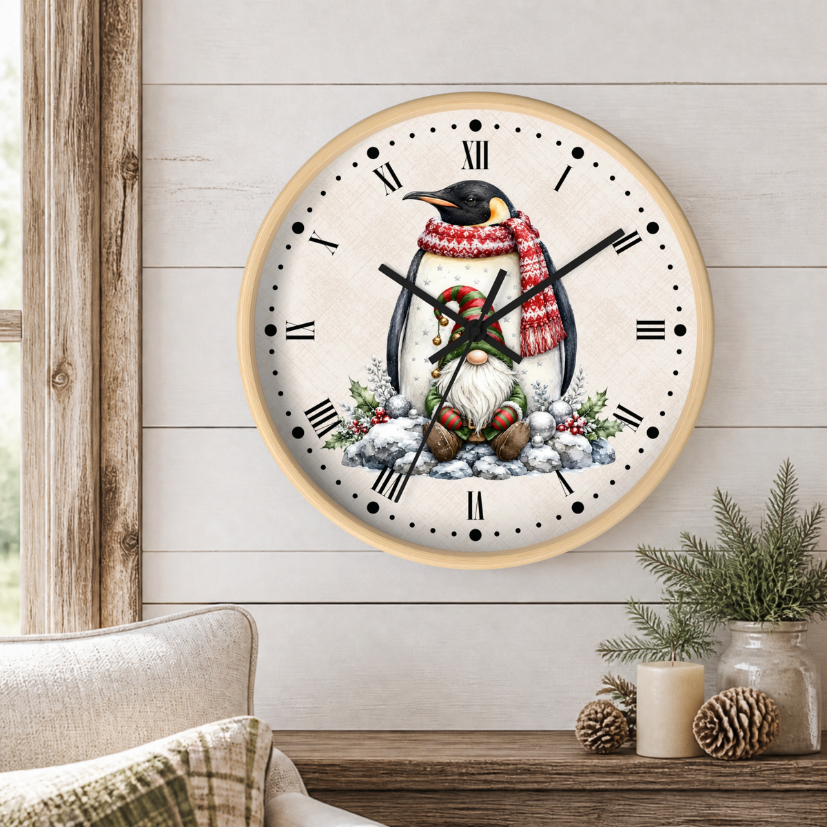 A round wall clock with a light wood frame hangs on a white shiplap wall above a rustic wooden table. The clock face features an illustration of a penguin wearing a red and white striped knit sweater and matching knit hat, sitting in the snow with a small gnome beside it.