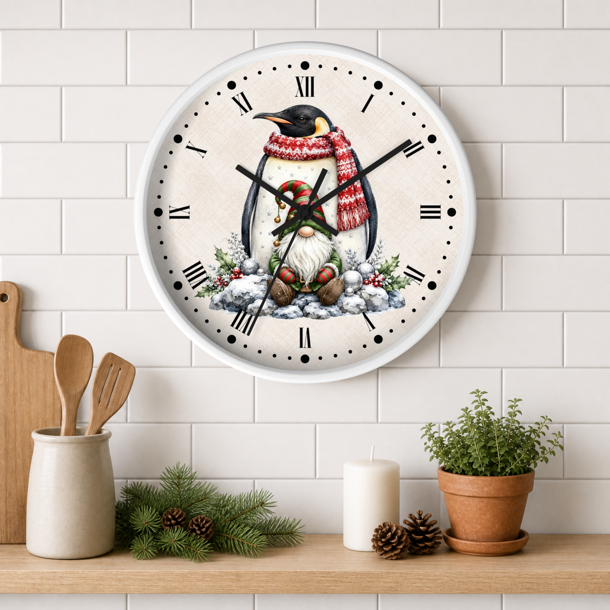 A round wall clock with a white frame hangs on a white subway-tile wall above a wooden shelf. The clock face features a whimsical illustration of a penguin wearing a red and white striped knit scarf and a green hat, surrounded by snow and evergreen branches.
