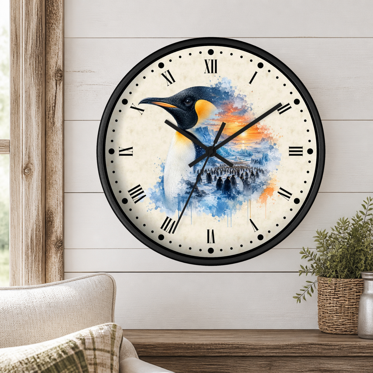 A round wall clock with a black frame and a penguin illustration over blue water and orange sky, mounted on a white shiplap wall above a wooden table with a plant and part of a sofa visible.