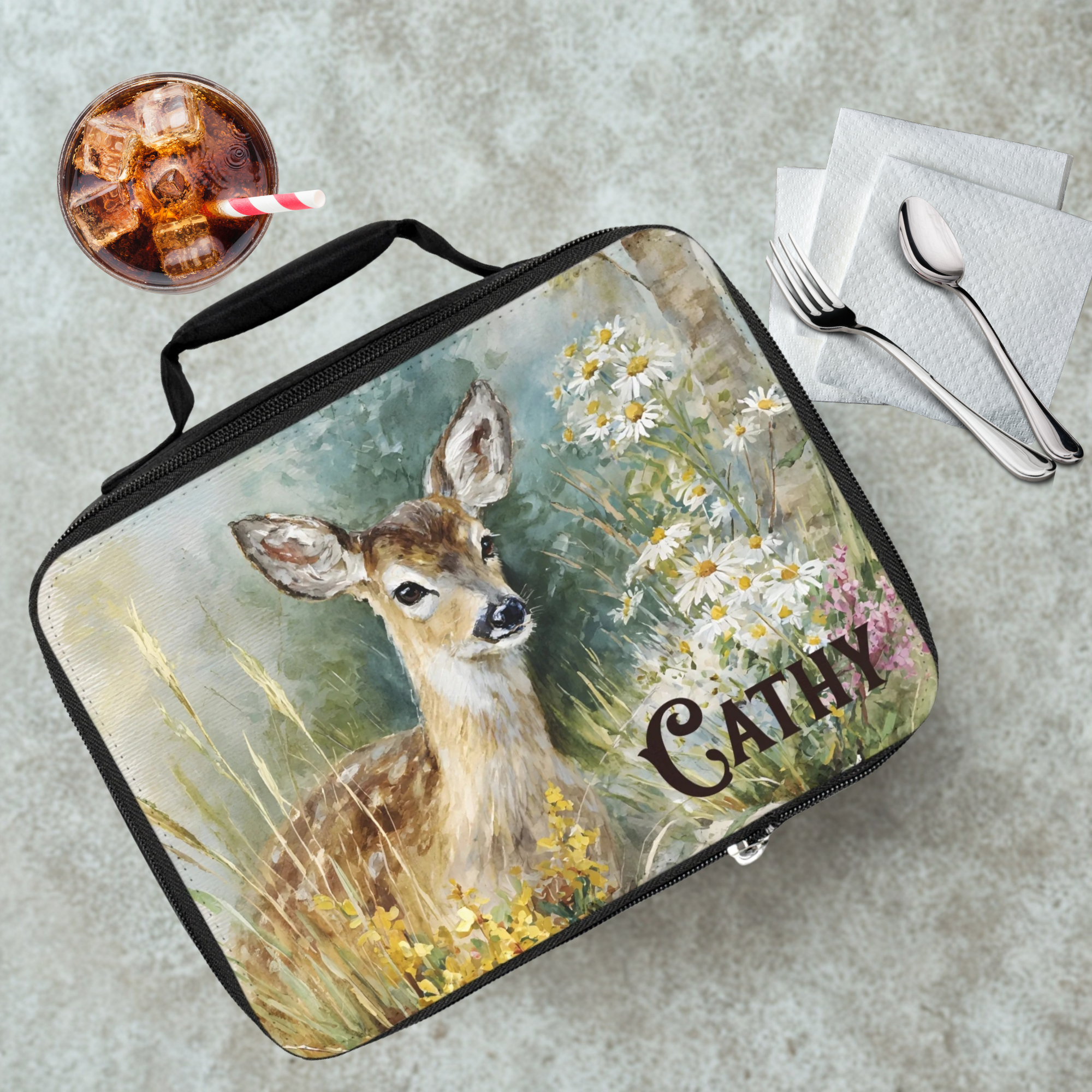  insulated lunch bag with a deer design and the name 'Cathy' on a cafeteria table with a drink and utensils.
