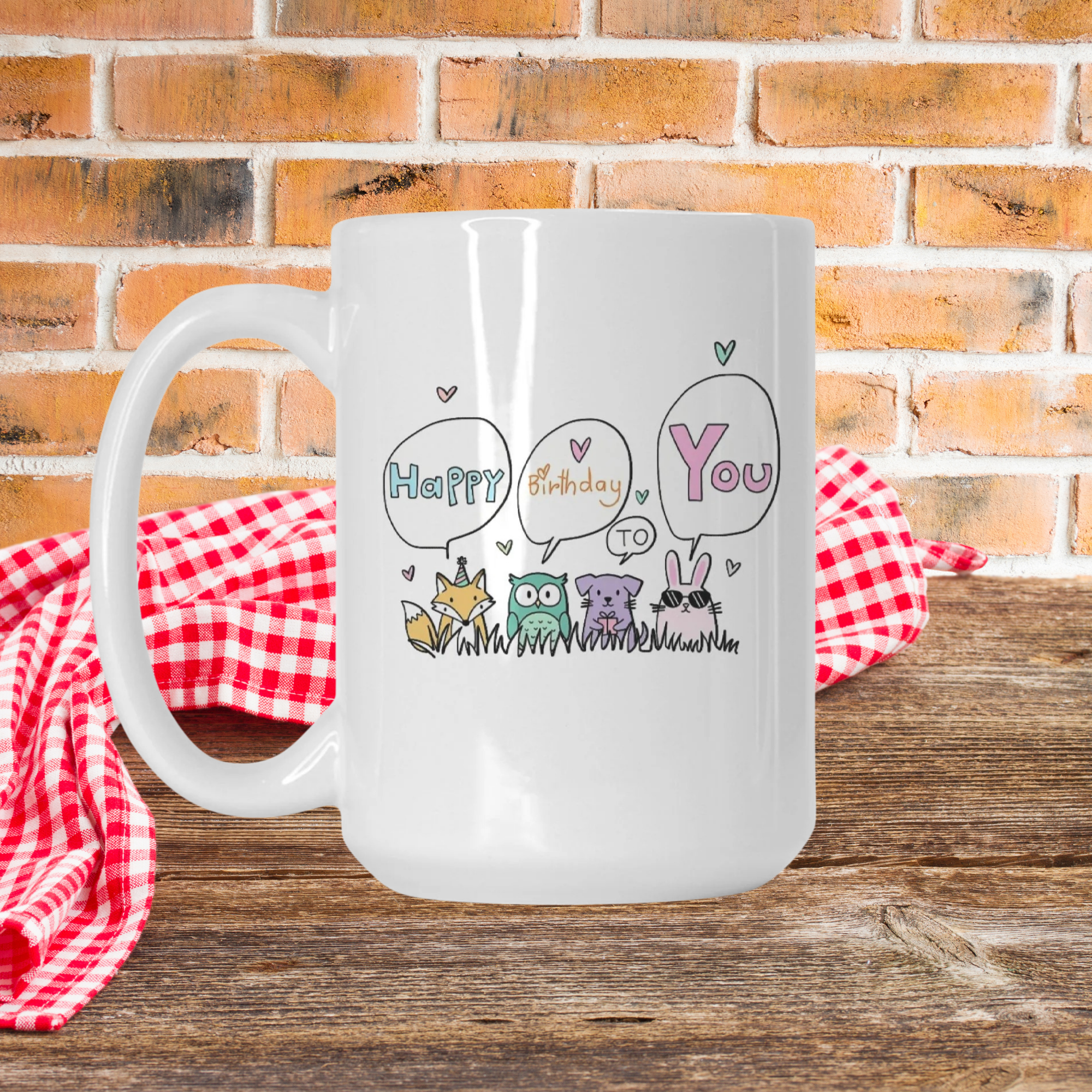 White mug with 'Happy Birthday to You' design on a wooden surface with a red checkered cloth and brick wall background.