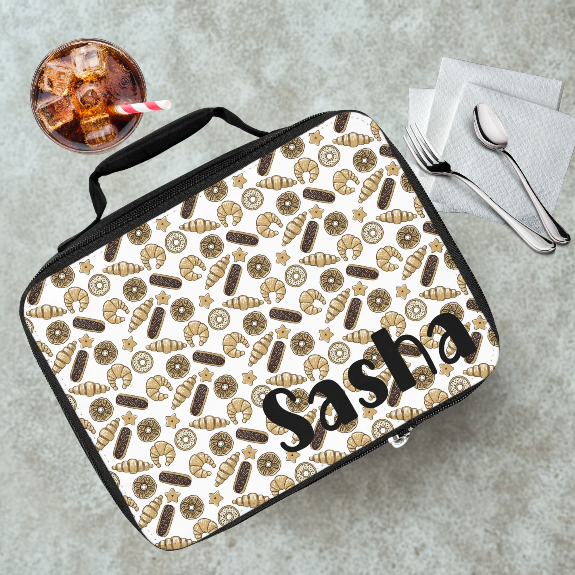 Lunch bag with baked goods pattern and 'Sasha' text on a cafeteria table with a drink and utensils.