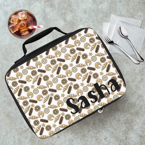 Lunch bag with baked goods pattern and 'Sasha' text on a cafeteria table with a drink and utensils.