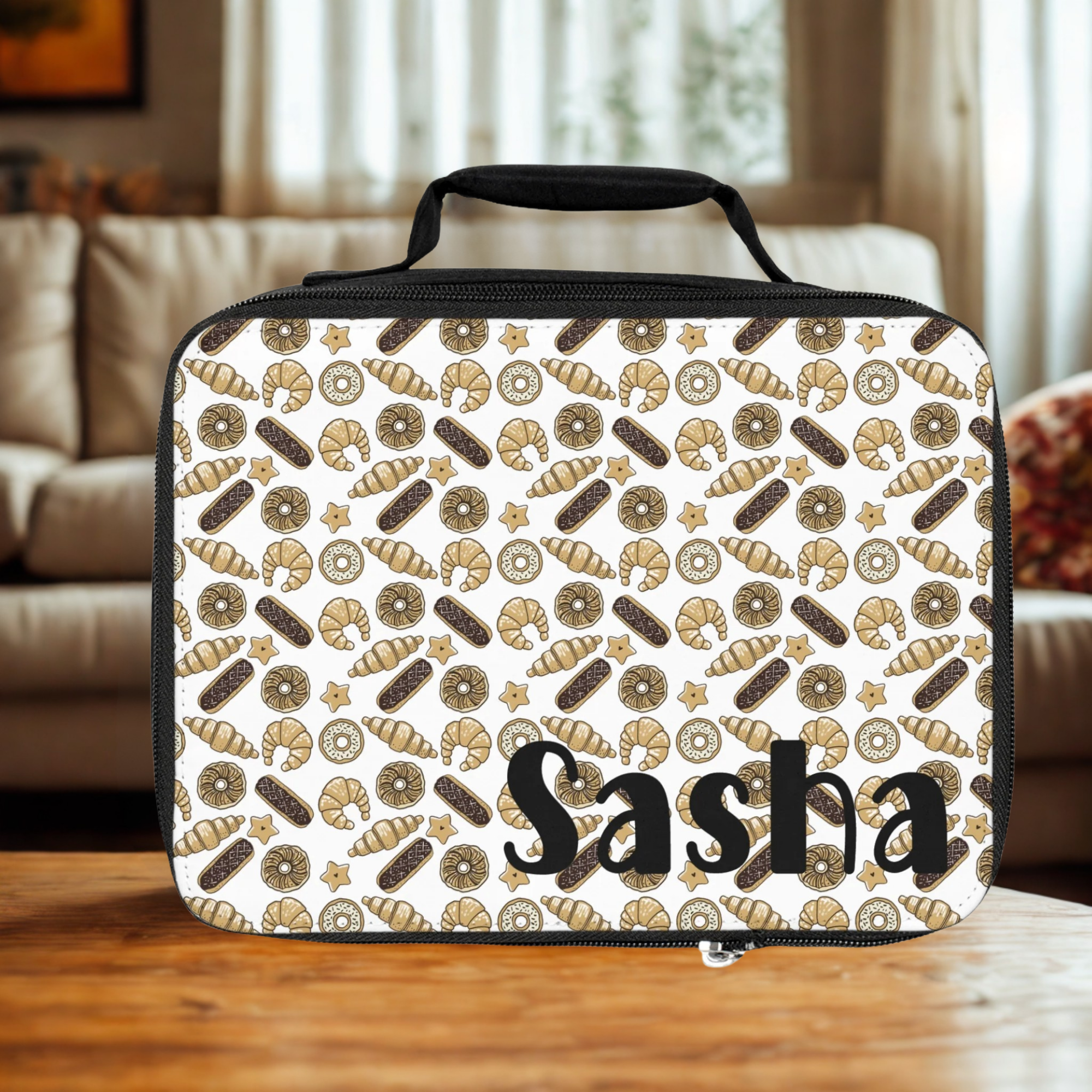 Lunch bag with baked goods pattern and 'Sasha' text on a wooden table in a living room.