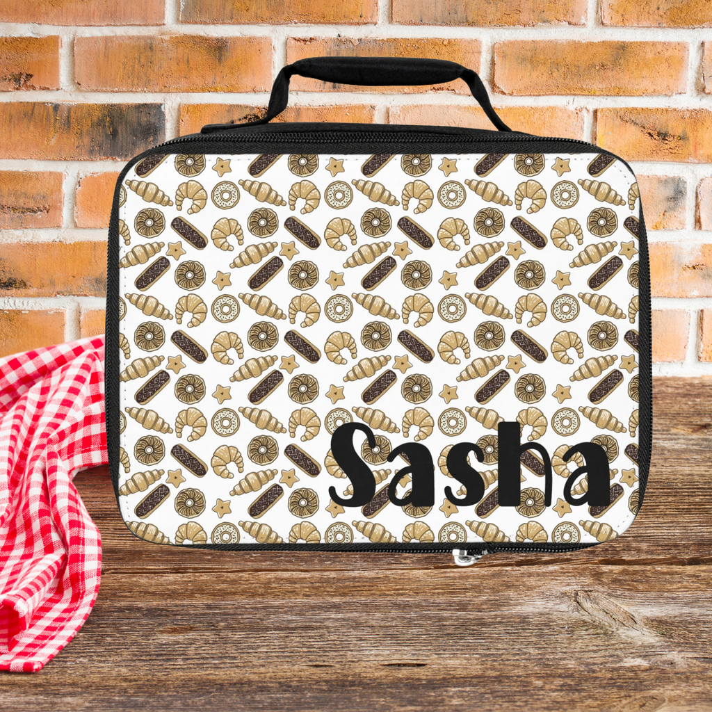 Lunch bag with donut, croissant, and long john pattern and 'Sasha' text on a wooden picnic table with brick wall background