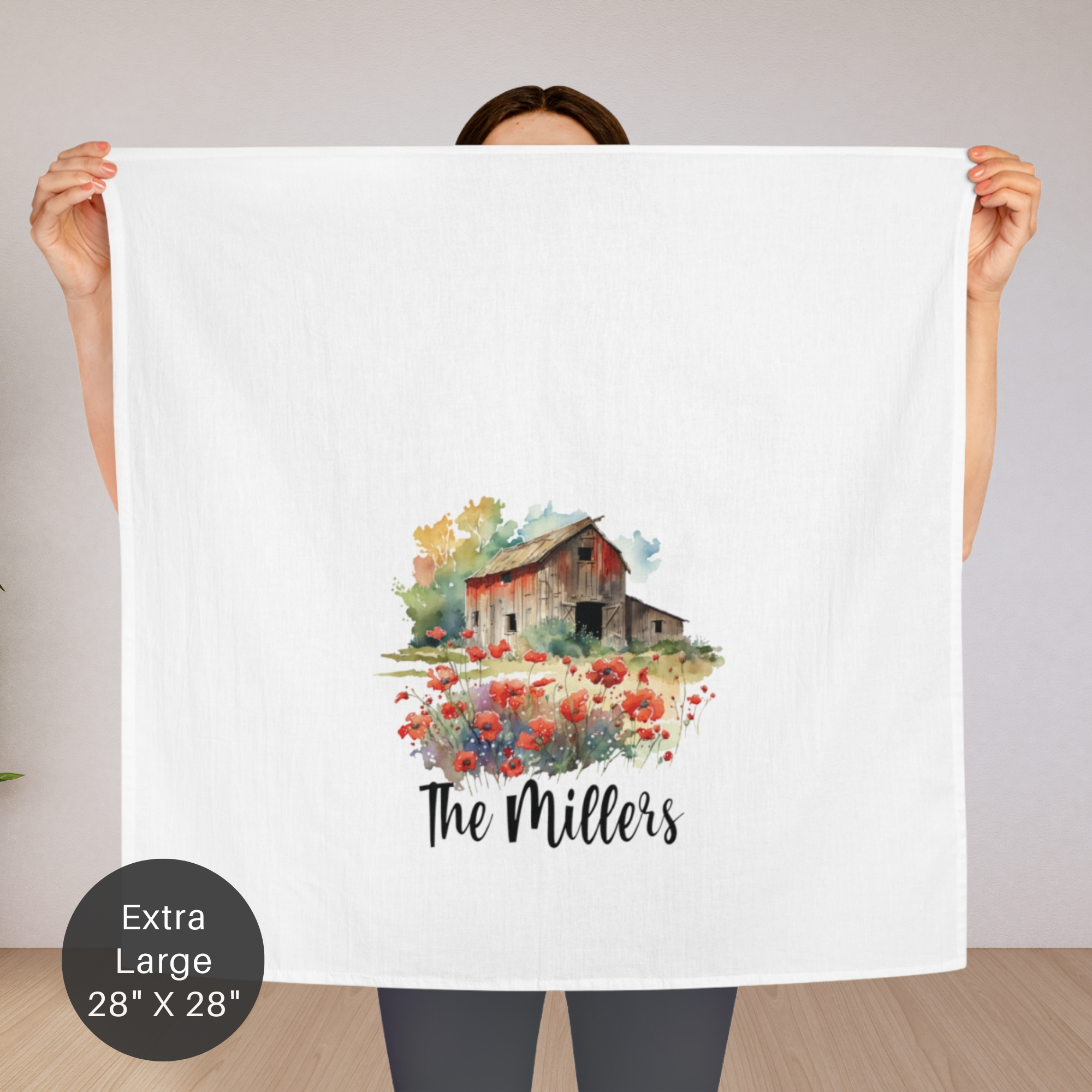Person holding an extra large tea towel with a barn and flowers design and 'The Millers' text.