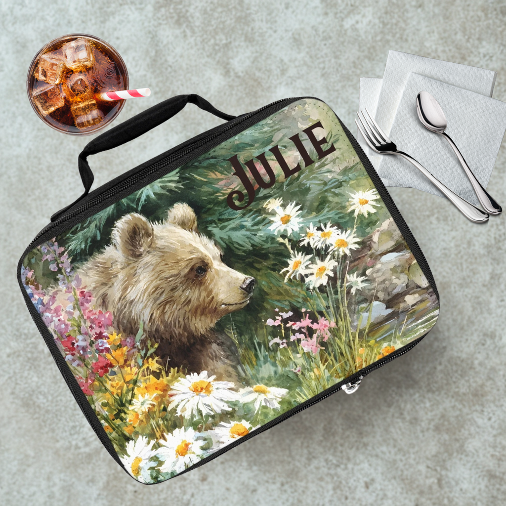 Lunch bag with bear design and 'Julie' text on a cafeteria table with a drink and utensils.