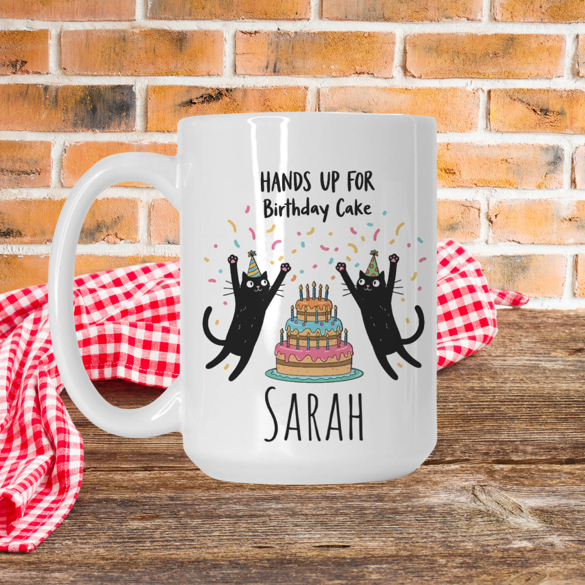 White mug with black cats and birthday cake design on a wooden picnic table with a brick wall background