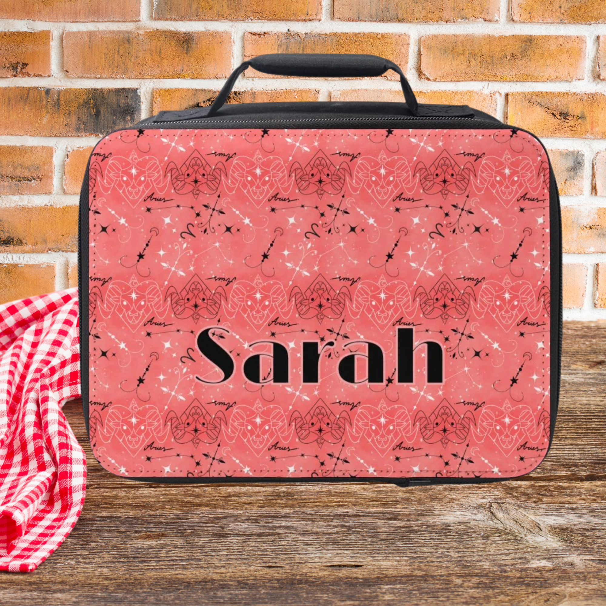 Pink patterned lunch bag with 'Sarah' printed on it, placed on a wooden surface with a brick wall background.