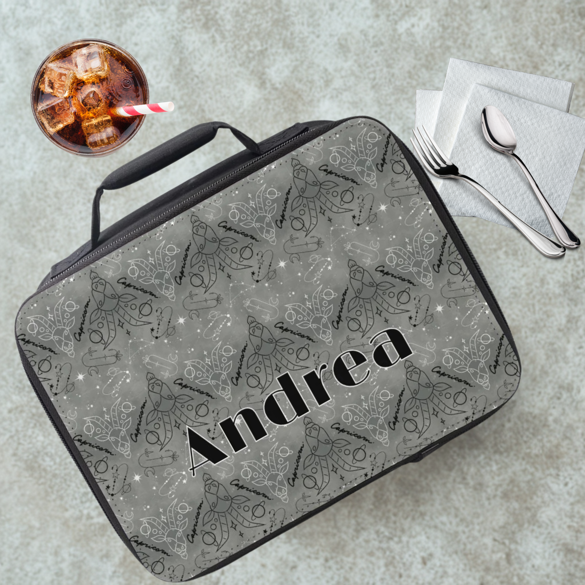 Gray insulated lunch bag with 'Andrea' custom text on a cafeteria table with a drink and utensils.