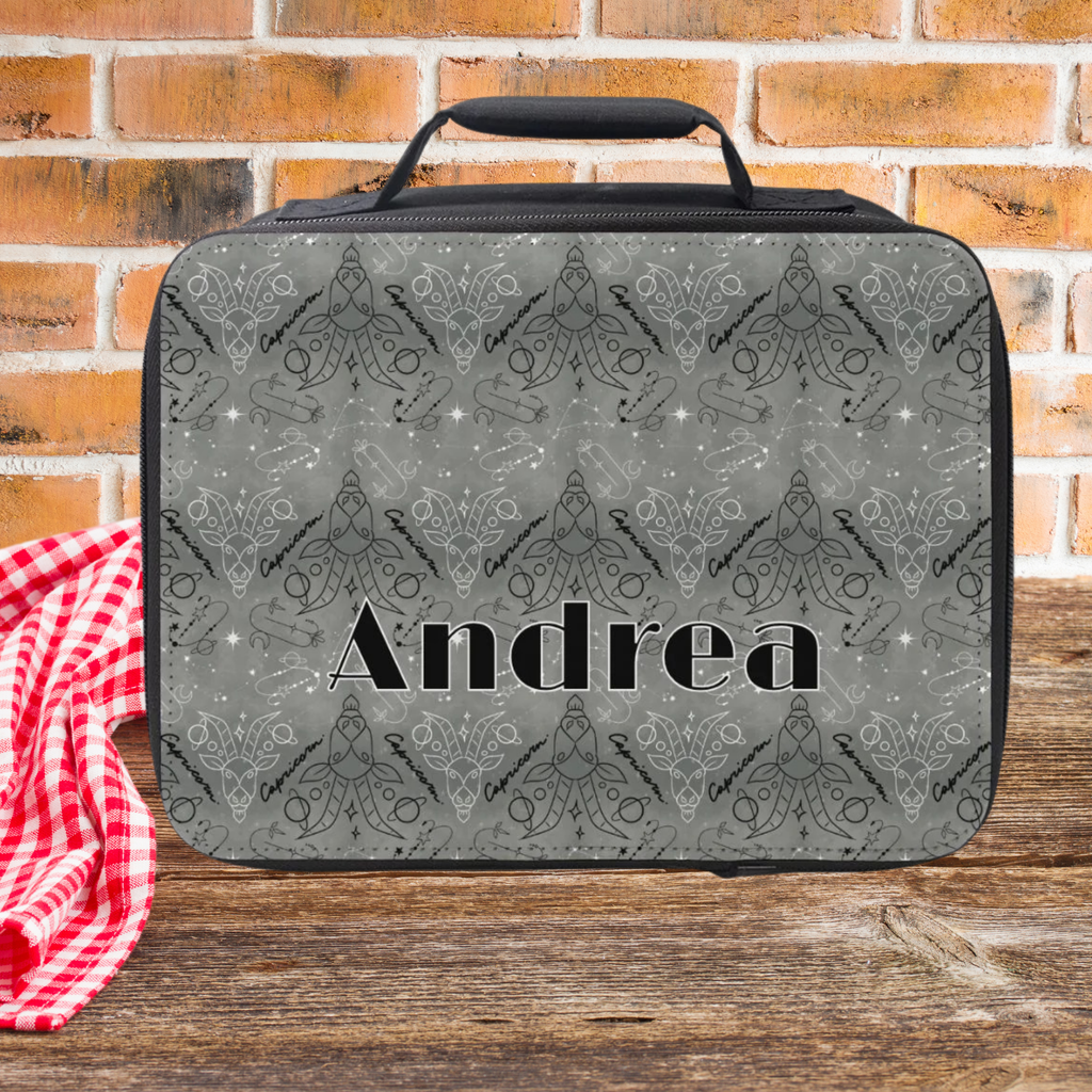 Lunch bag with patterned Capricorn zodiac design and 'Andrea' text on a wooden picnic table against a brick wall background