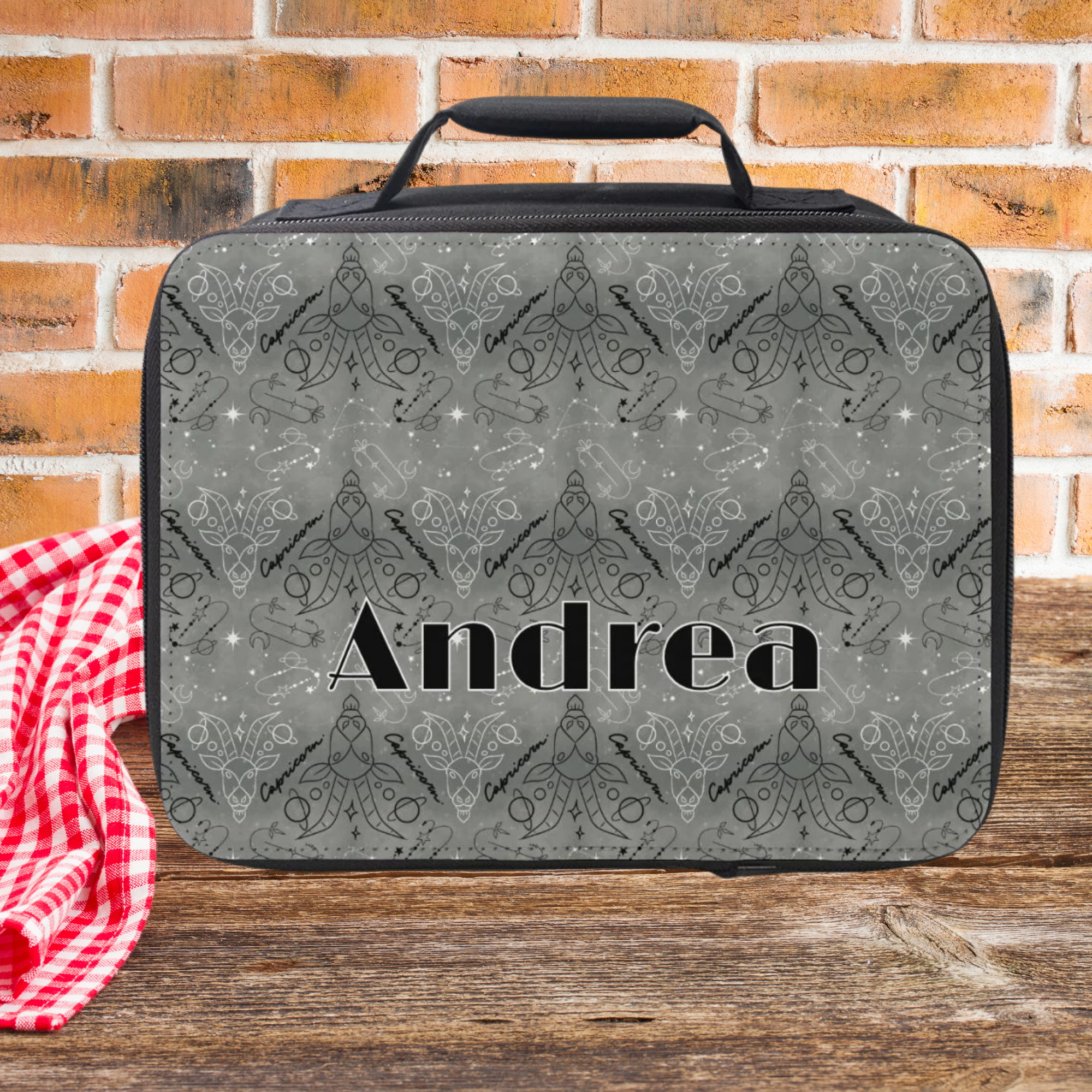 Lunch bag with patterned Capricorn zodiac design and 'Andrea' text on a wooden picnic table against a brick wall background