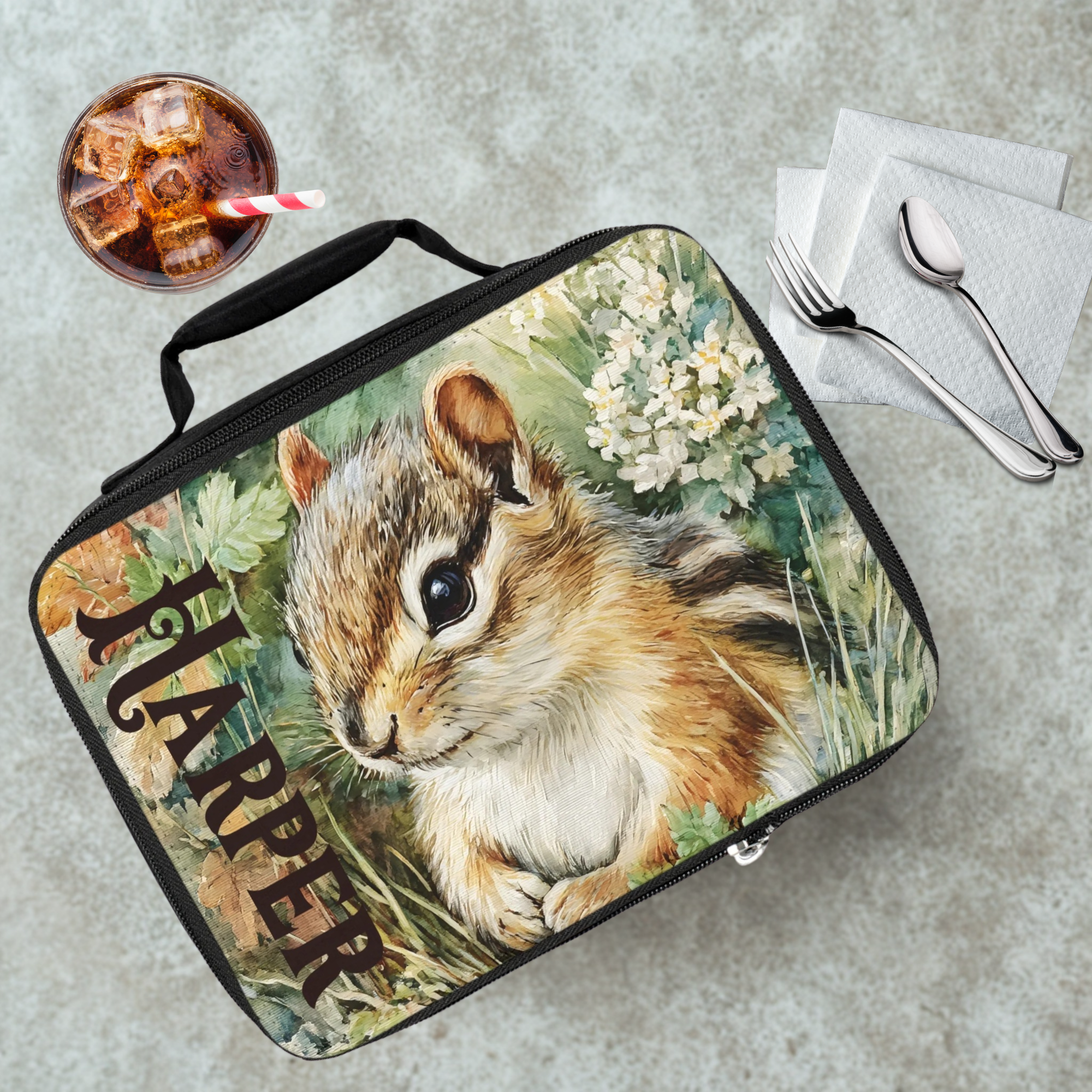 Lunch bag with a chipmunk design and the name 'Harper' on a gray tab;e with a drink and utensils.
