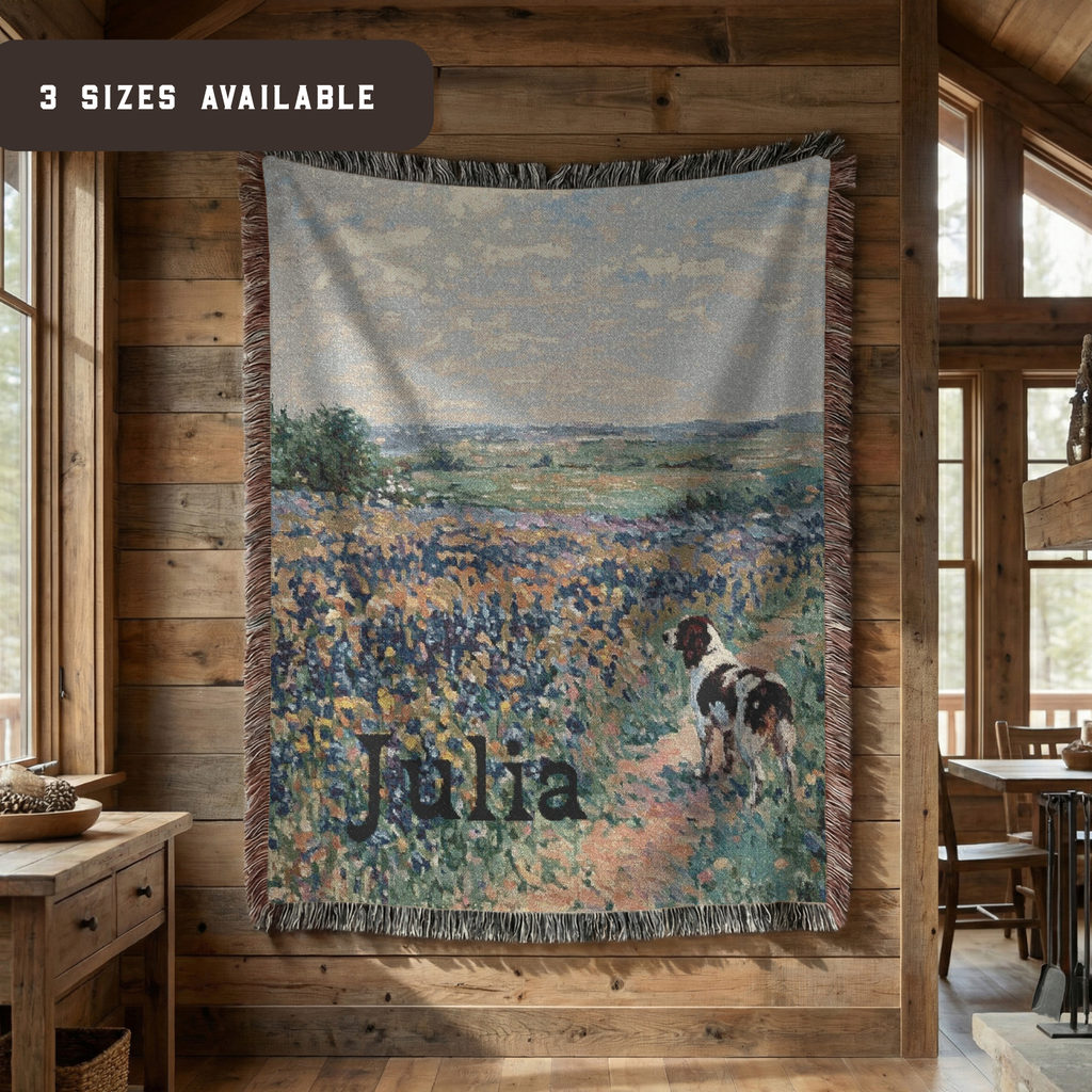 A personalized woven throw blanket featuring an impressionistic blue flower field with a dog and the name 'Julia', hanging on a rustic wood-paneled wall in a cabin interior. A banner in the top left reads '3 SIZES AVAILABLE'.