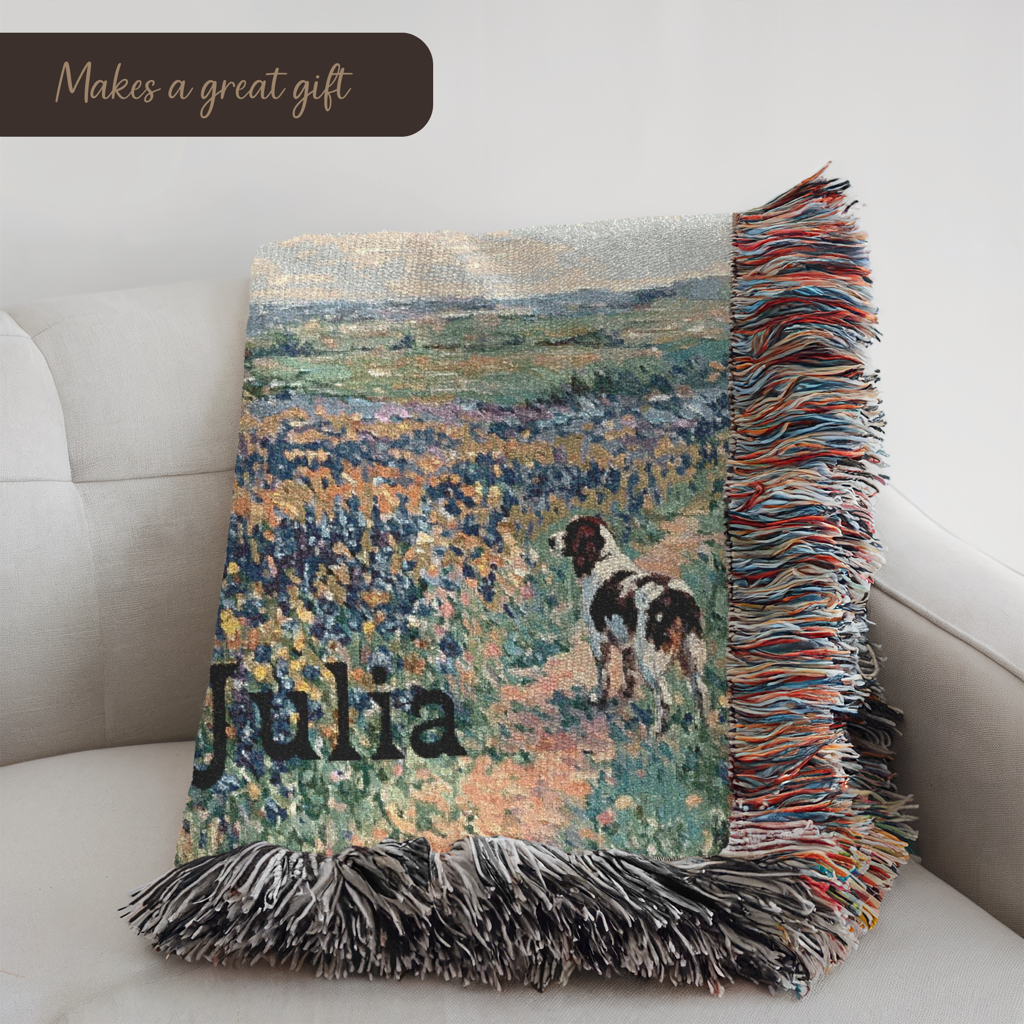 A woven throw blanket draped over a light beige sofa, featuring a painted landscape of a dog in a field, the name 'Julia' in black script, and multicolored fringe on all sides.