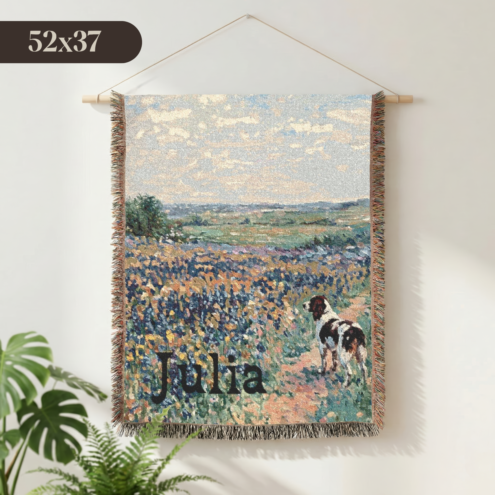 A personalized woven wall tapestry featuring a landscape painting of a cocker spaniel dog in a flower field, hanging on a white wall with a plant in the foreground. The tapestry has a wooden dowel and string hanger, and the name 'Julia' is printed at the bottom.