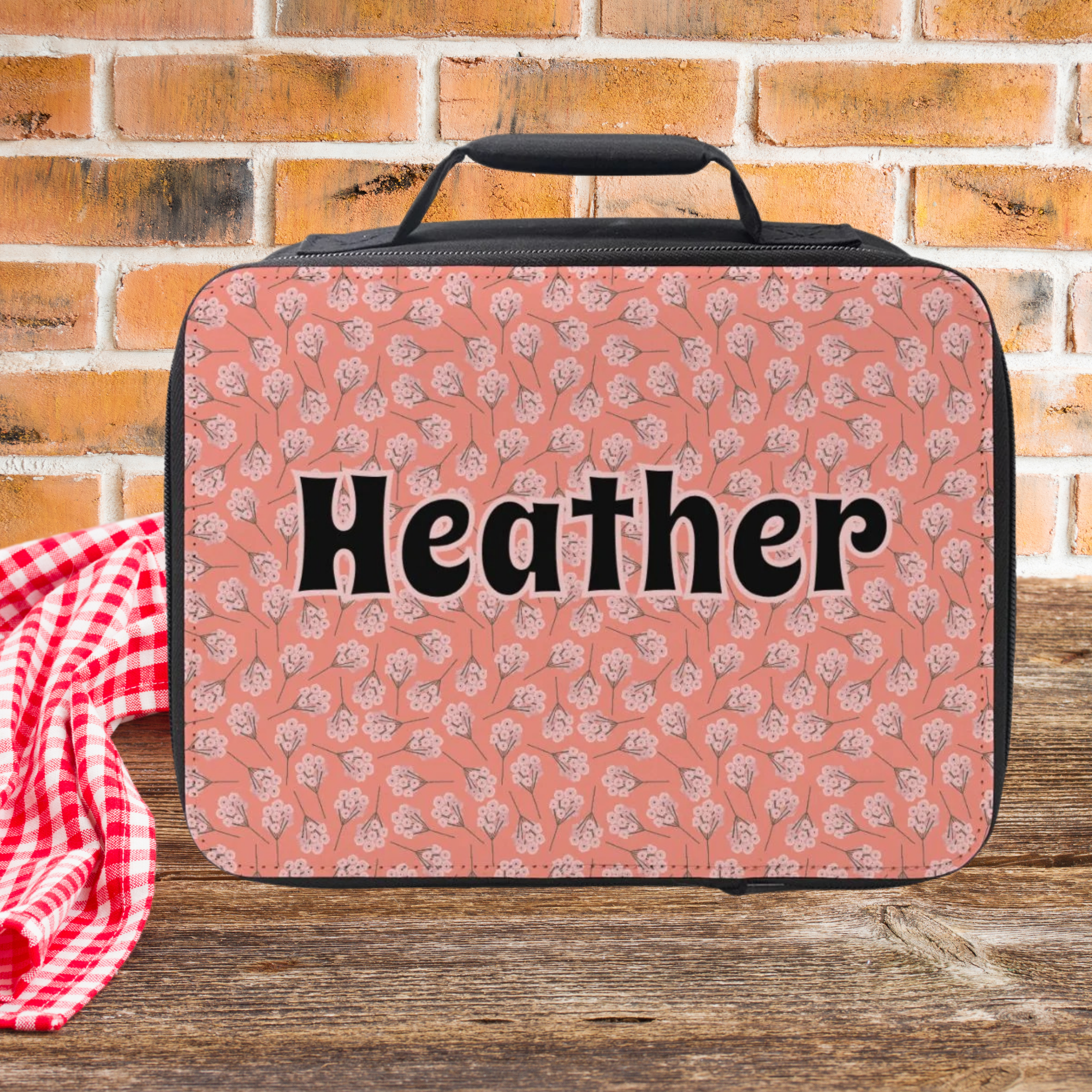 Pink floral-patterned lunch bag with 'Heather' on it against a brick wall background