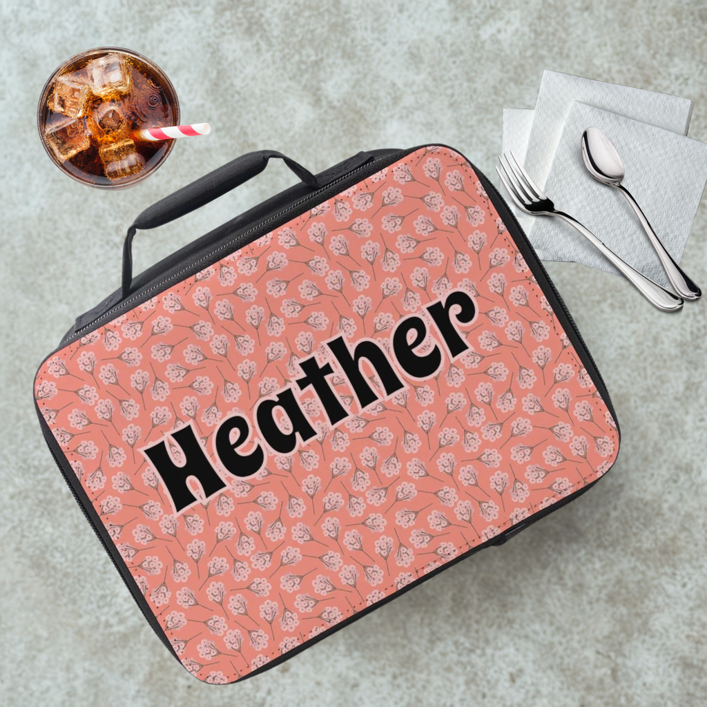 Pink floral-patterned lunch bag with 'Heather' printed on it, placed on a gray surface with a drink and utensils.