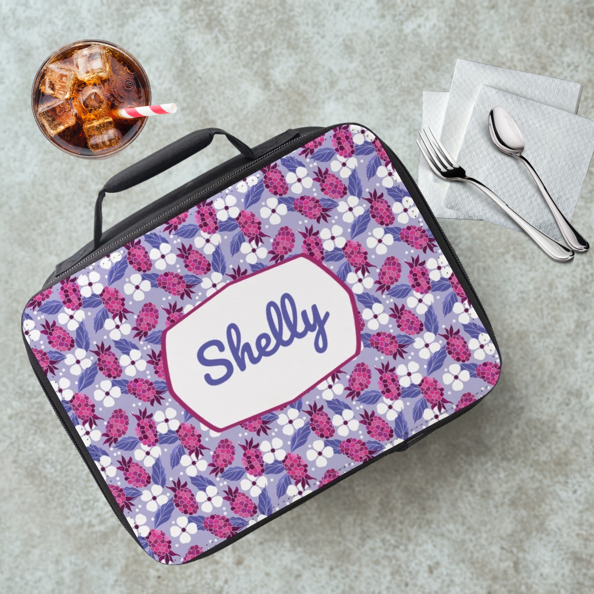 Floral-patterned lunch bag with 'Shelly' monogram on a cafeteria table with a drink and utensils.