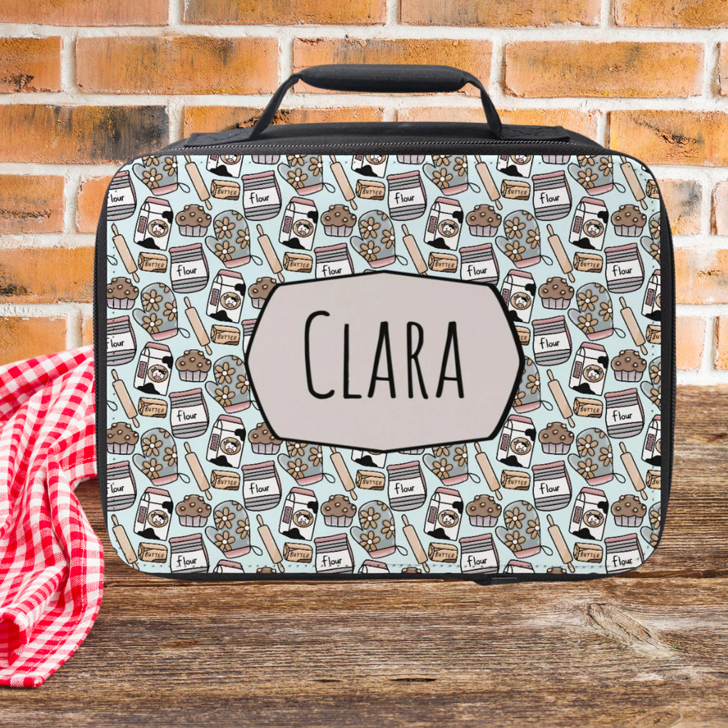 Lunch bag with baking pattern design and 'CLARA' personalized name  on a wooden picnic table with a brick wall background