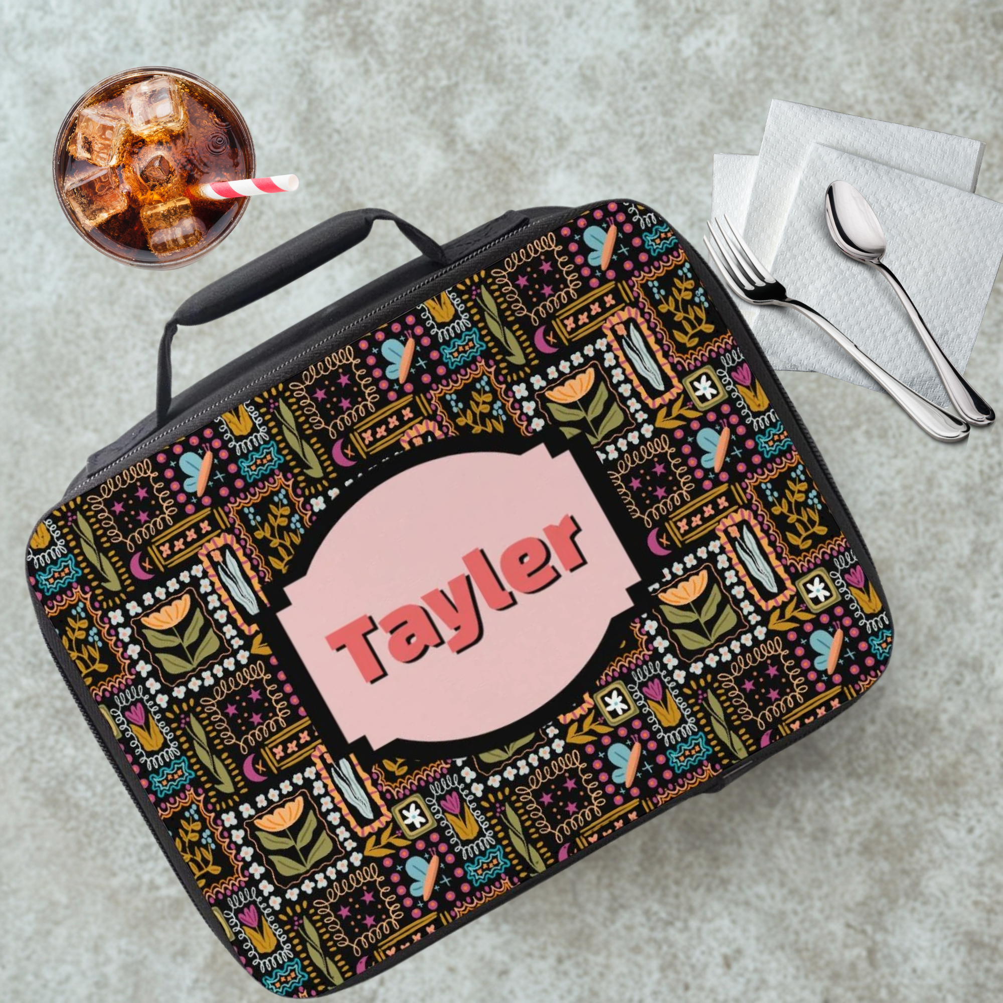 Lunch bag with colorful pattern and 'Tayler' name on a cafeteria table with a drink and utensils.