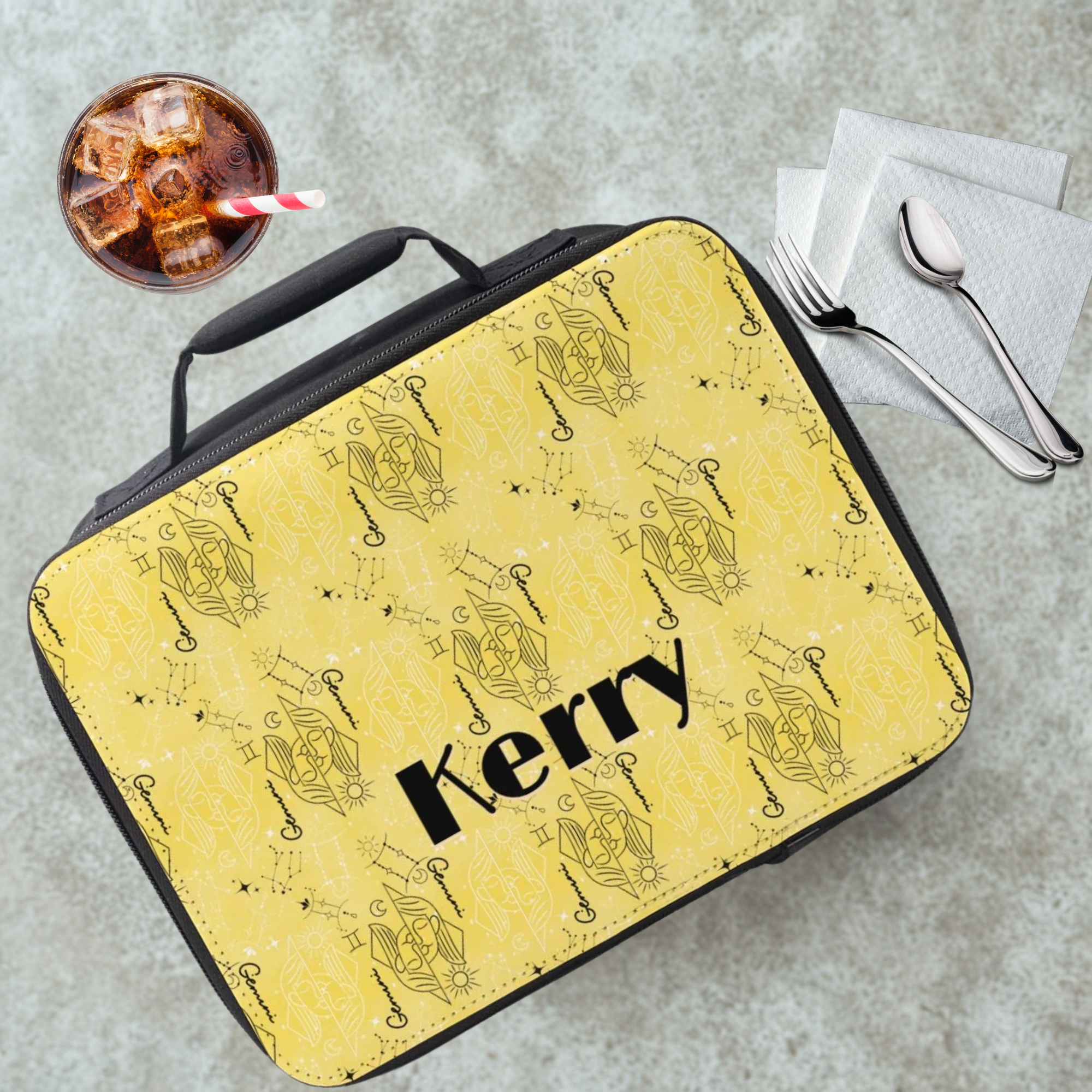 insulated lunch bag with yellow and black Gemini zodiac patterns and 'Kerry' custom text on a gray table with a drink and utensils.