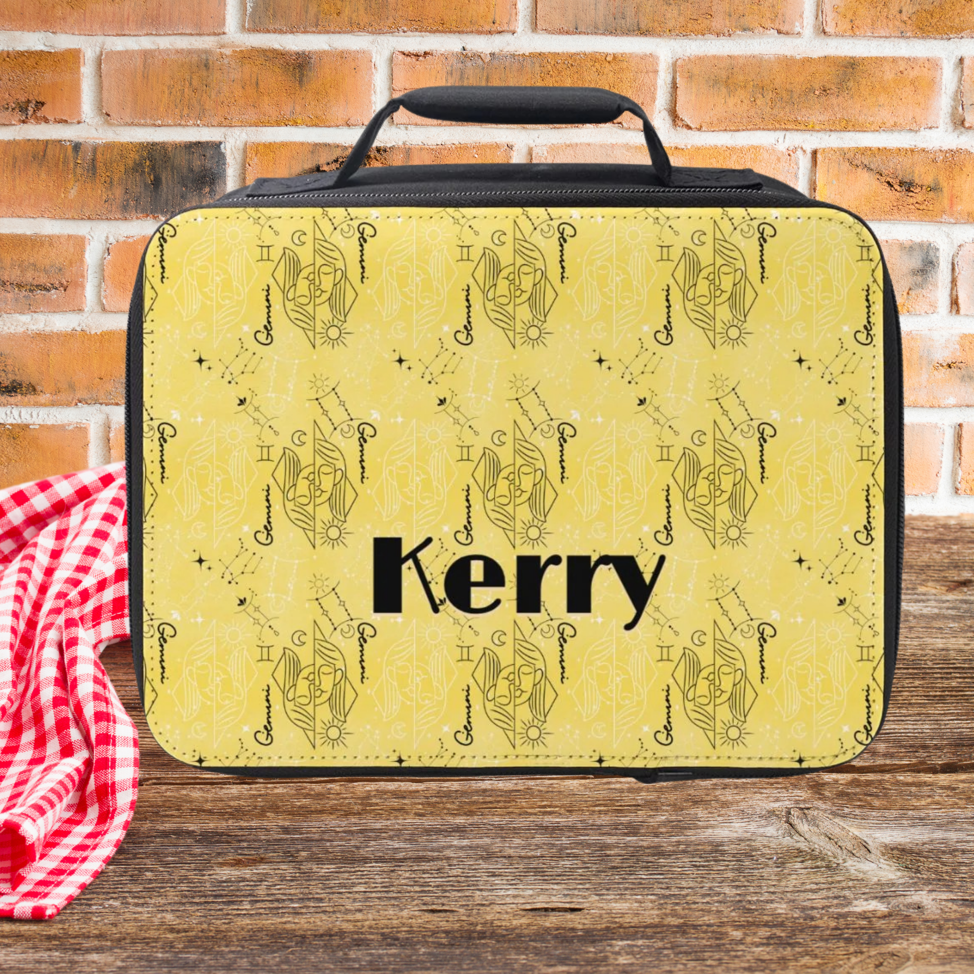 Yellow Gemini zodiac pattern lunch bag with 'Kerry' custom text on a wooden picnic table with a brick wall background