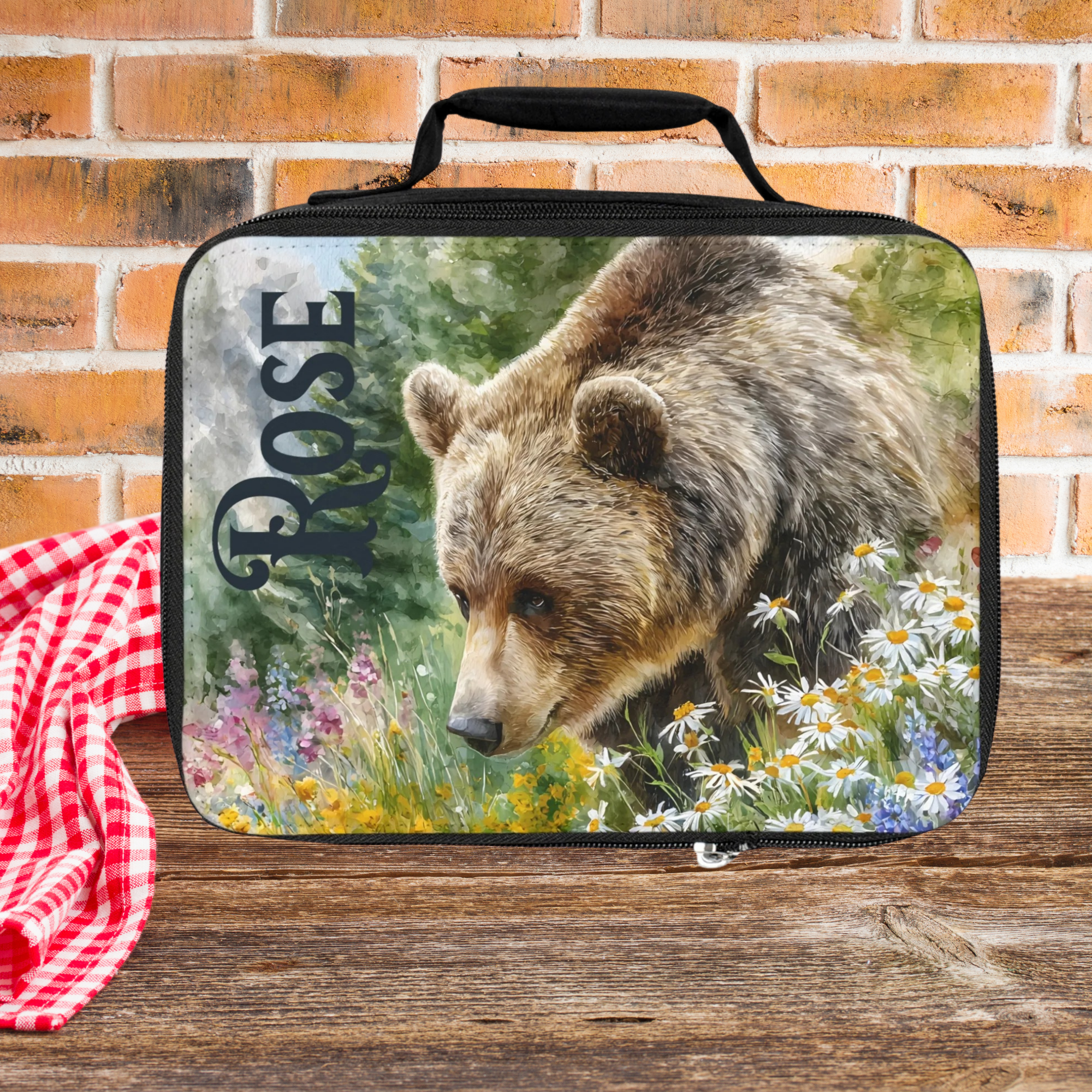 Lunch bag with bear design and 'Rose' custom text on a picnic table against a  brick wall background