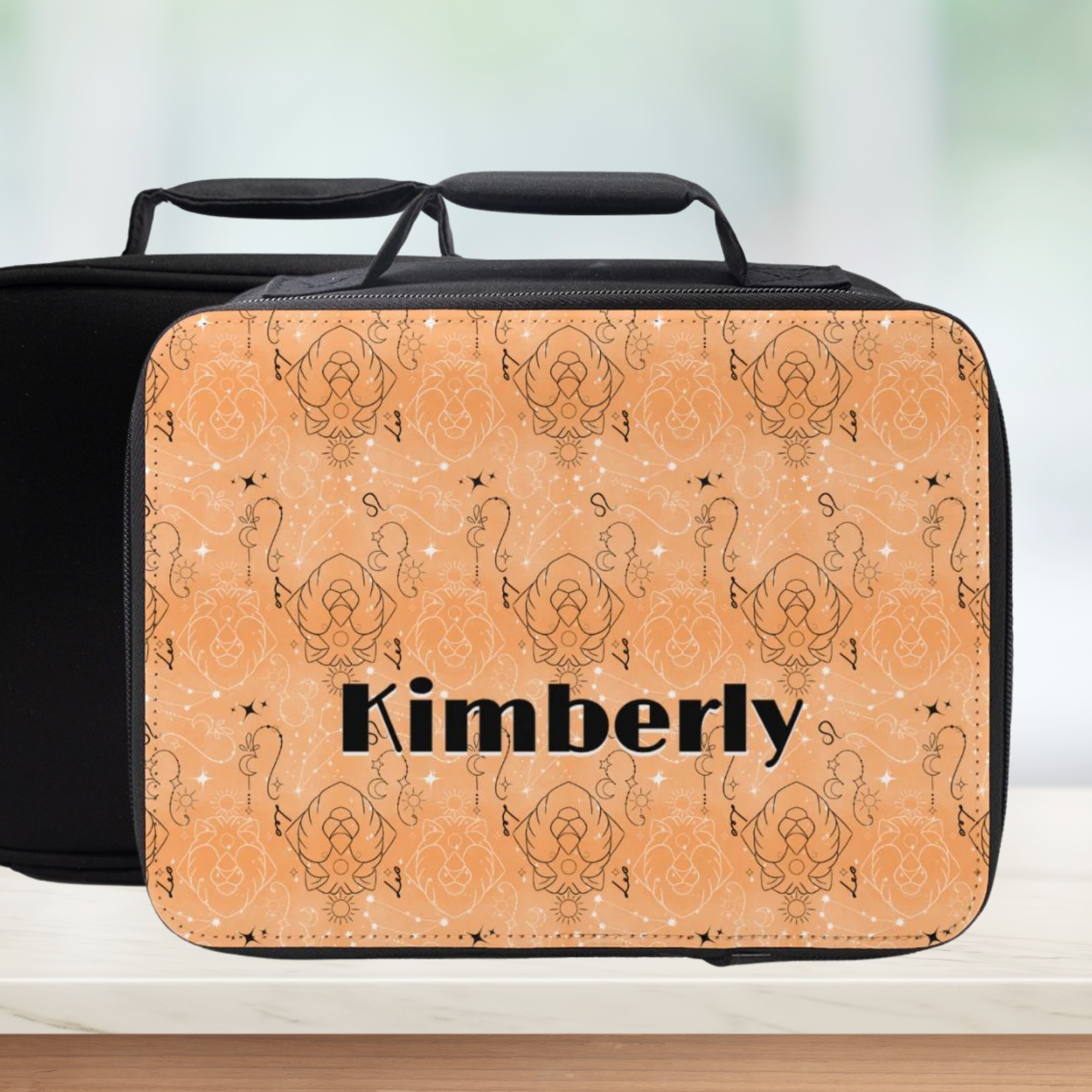 Leo zodiac patterned lunch bag with 'Kimberly' printed on it, black handle on a wood surface and blurred background.