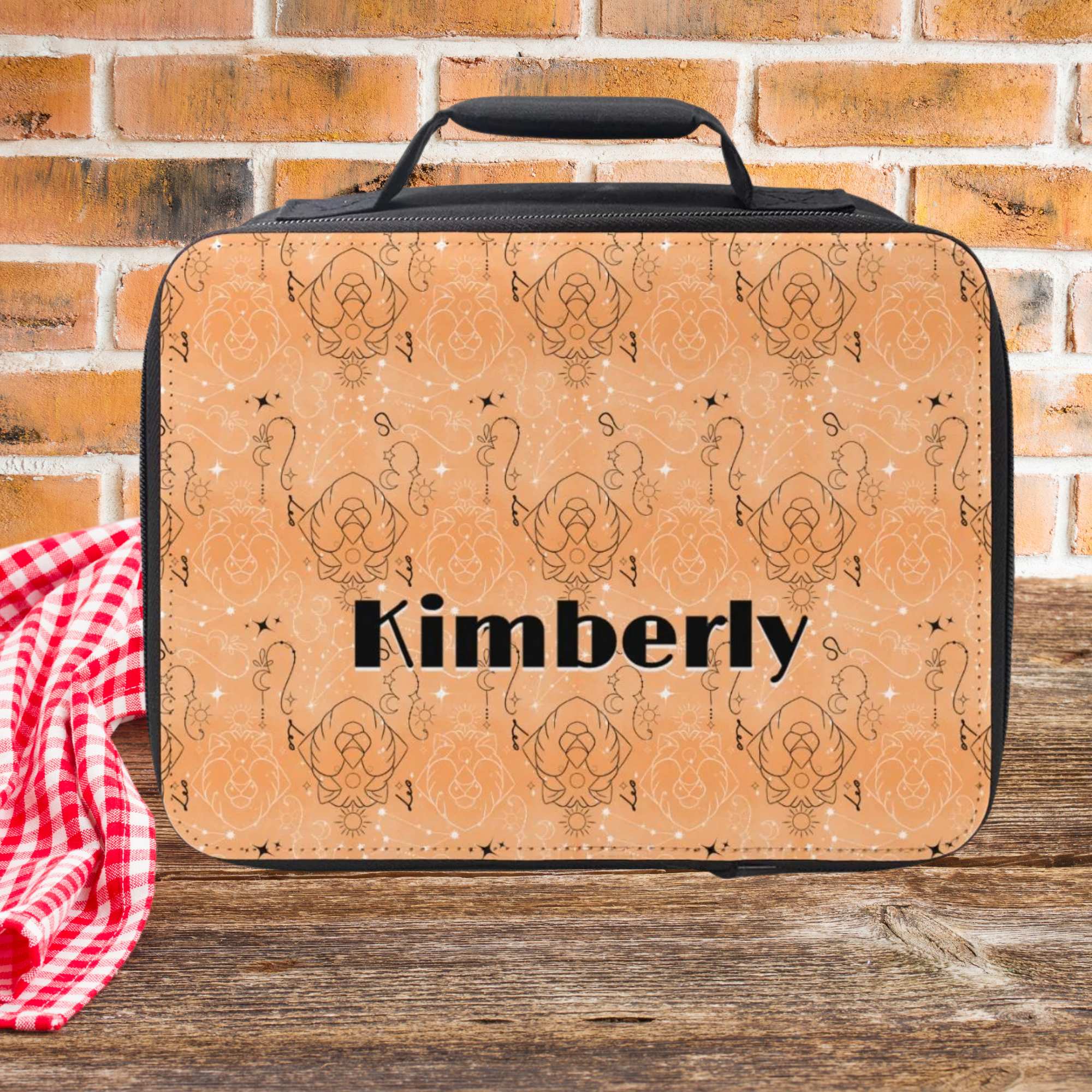 Leo zodiac patterned lunch bag with 'Kimberly' custom text on a wooden picnic table and brick wall background
