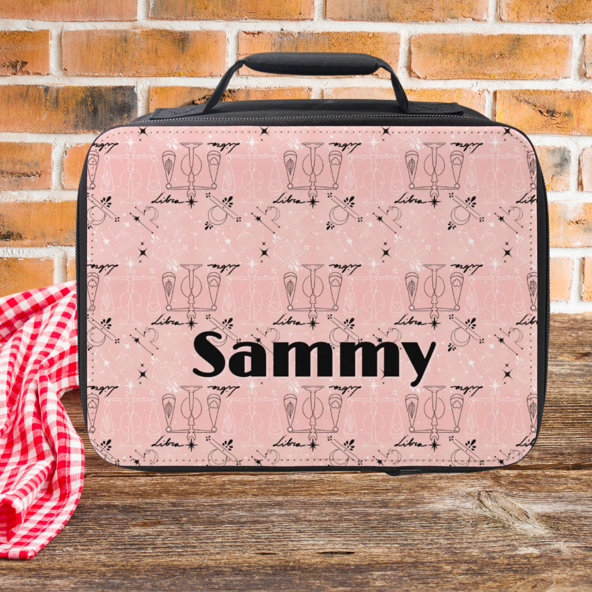 Pink and black Libra zodiac pattern lunch bag with 'Sammy' custom text on a wooden picnic table against a brick wall background