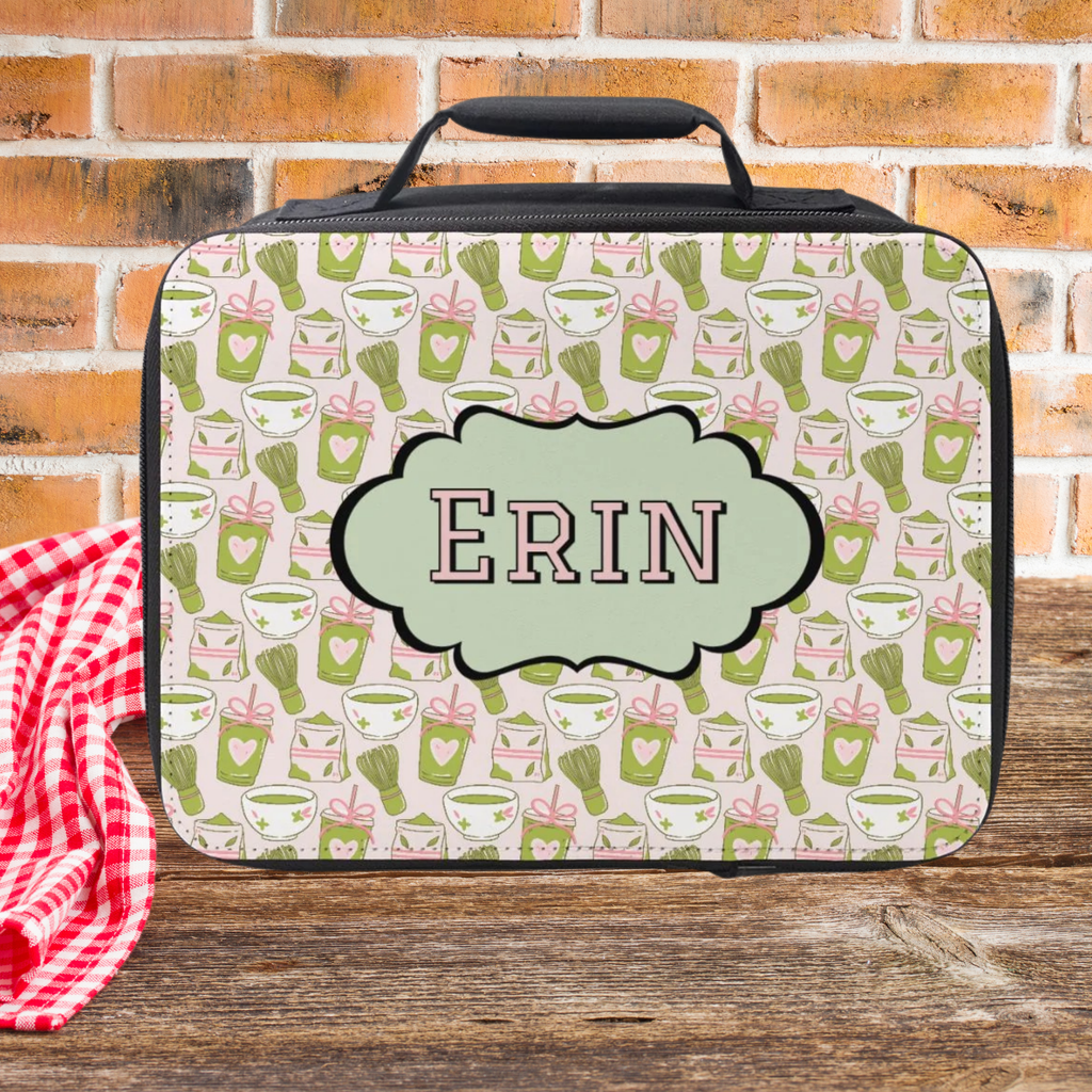 Personalized lunch bag with green matcha drink pattern and 'ERIN' monogram on a wooden picnic table with brick background