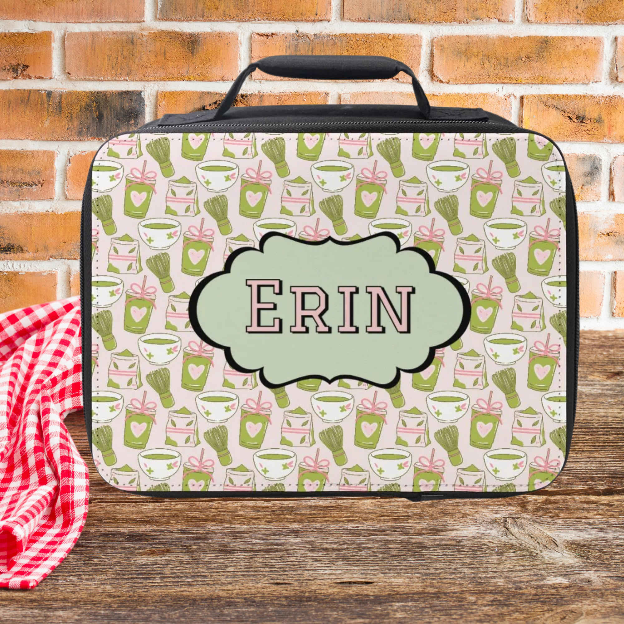 Personalized lunch bag with green matcha drink pattern and 'ERIN' monogram on a wooden picnic table with brick background