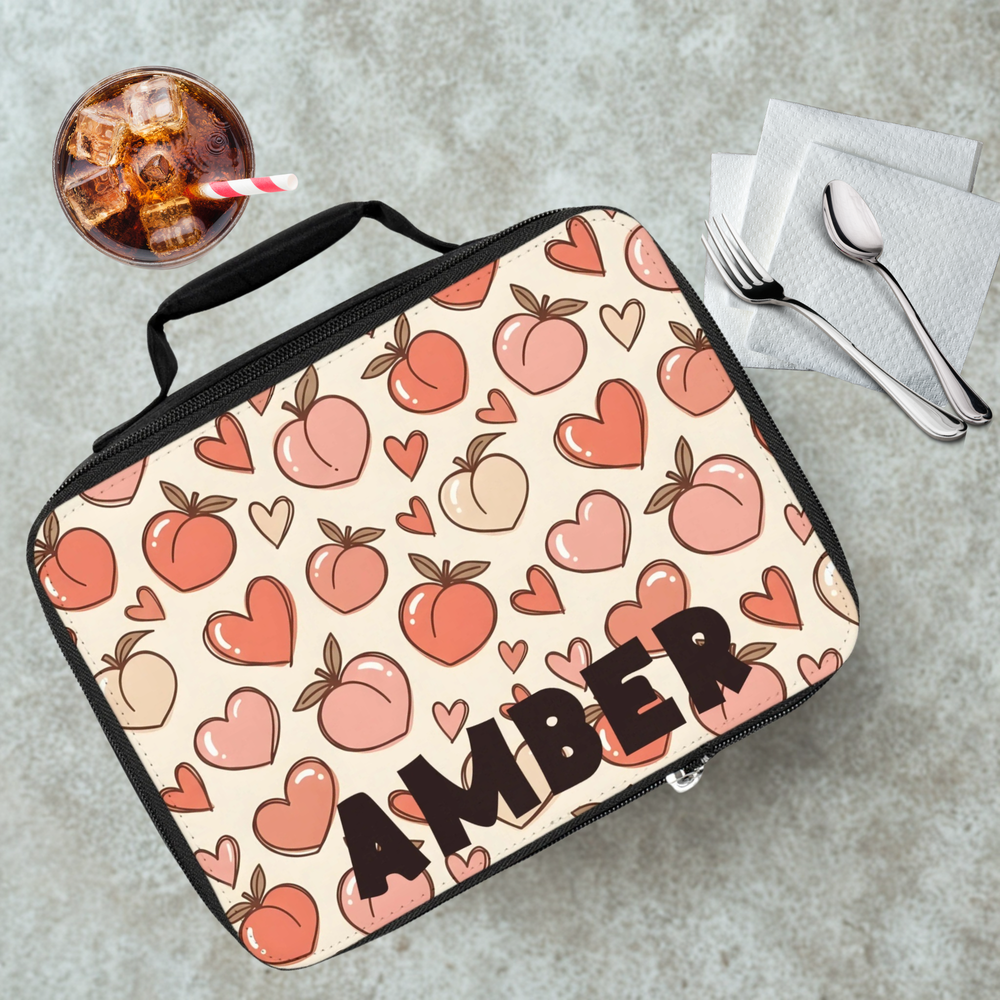 Lunch bag with peach and heart pattern and 'AMBER' personalized text on a cafeteria table with a drink and utensils.