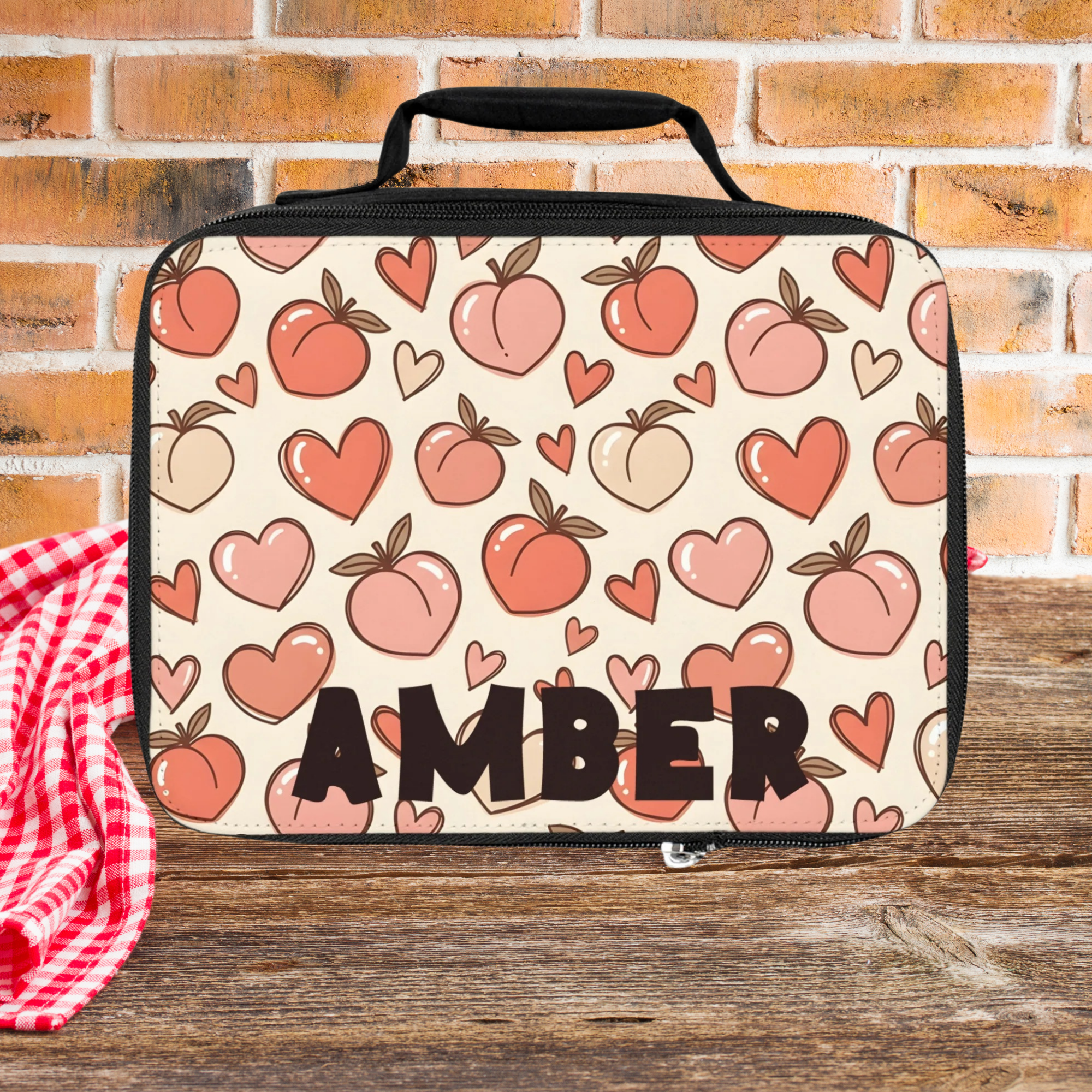 Lunch bag with peach and heart pattern and 'AMBER' personalized text on a picnic table against a brick wall background