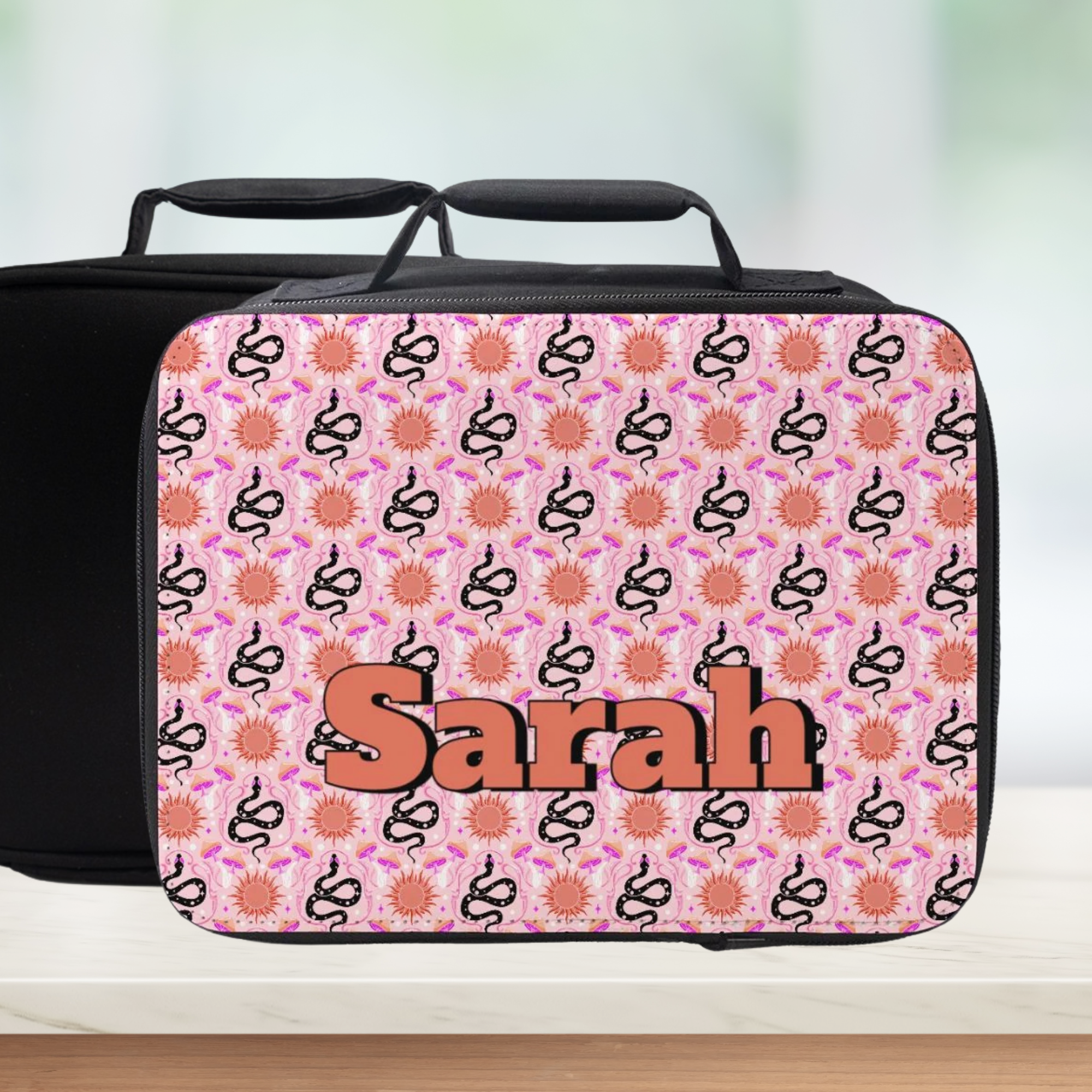 Pink lunch bag with black floral Celestial snake pattern and 'Sarah' custom text on a blurred background