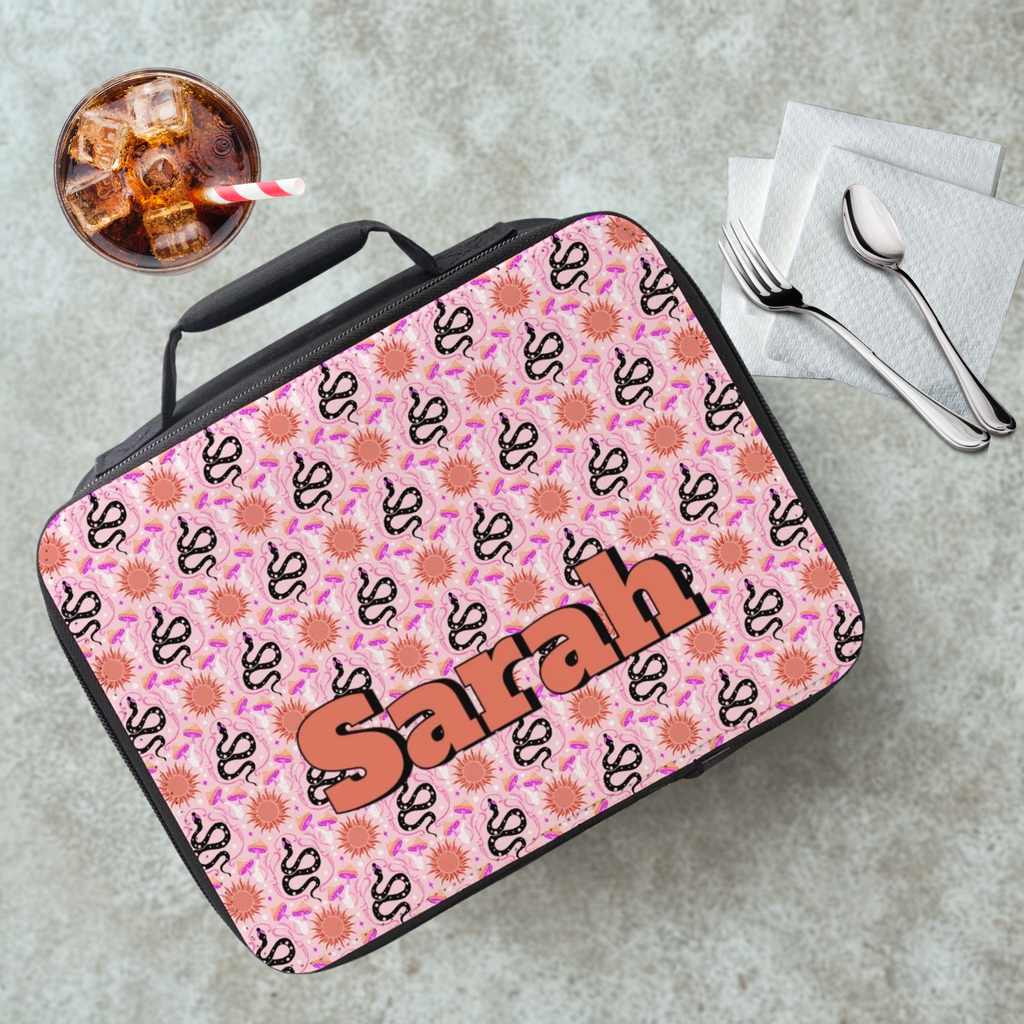 Pink patterned lunch bag with 'Sarah' printed on it, placed on a gray surface with a drink and utensils.