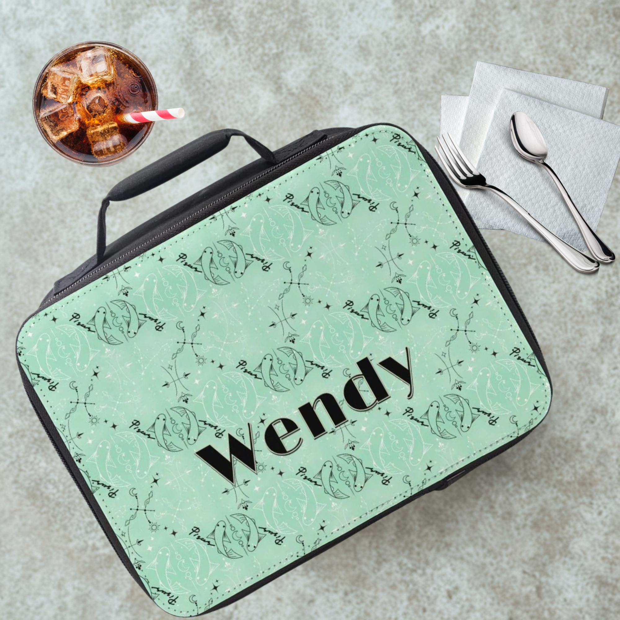 Green Pisces zodiac patterned lunch bag with 'Wendy' printed on it, placed on a cafeteria table with a drink and utensils in the background.