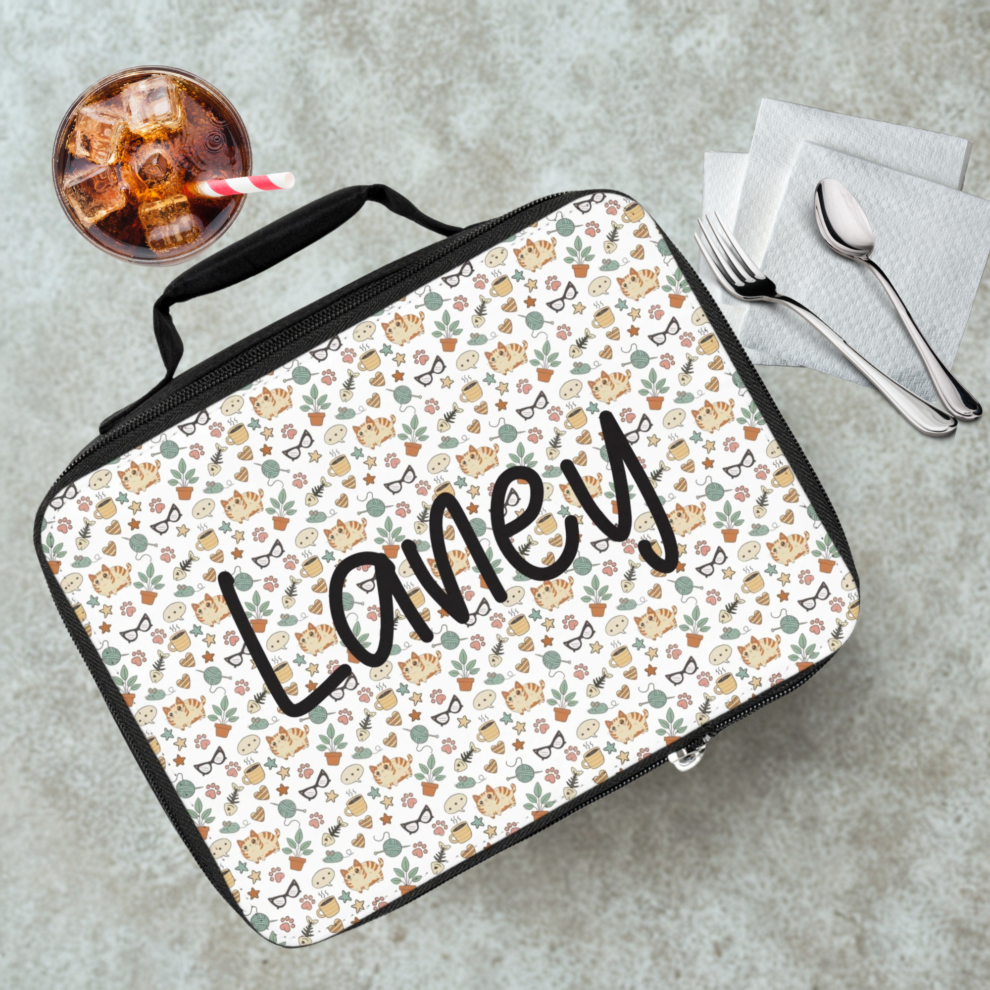 Lunch bag with cat  pattern and custom text on a cafeteria table with a drink and utensils.