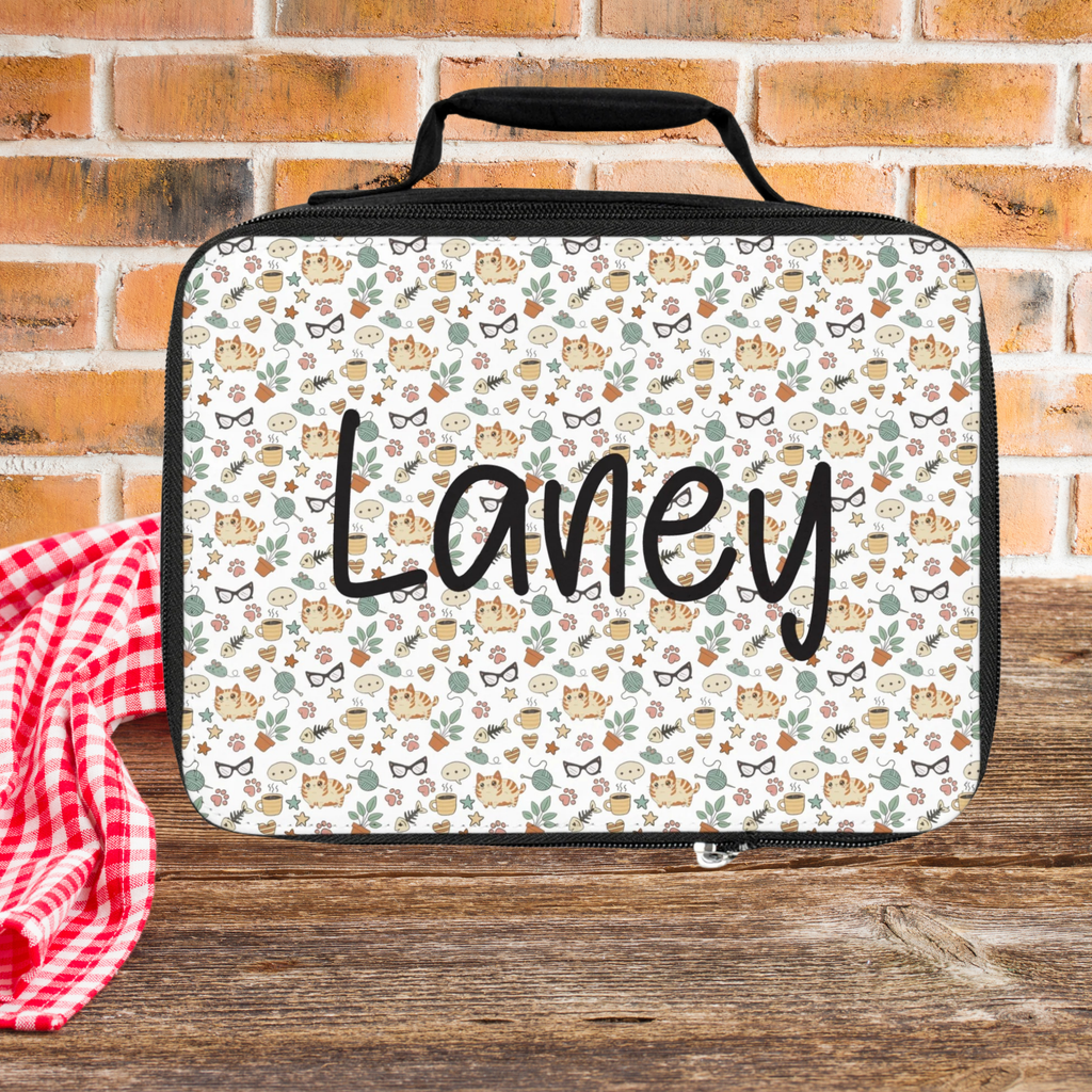 Lunch bag with a cat pattern and 'Laney' custom text on a wooden picnic table and brick wall background