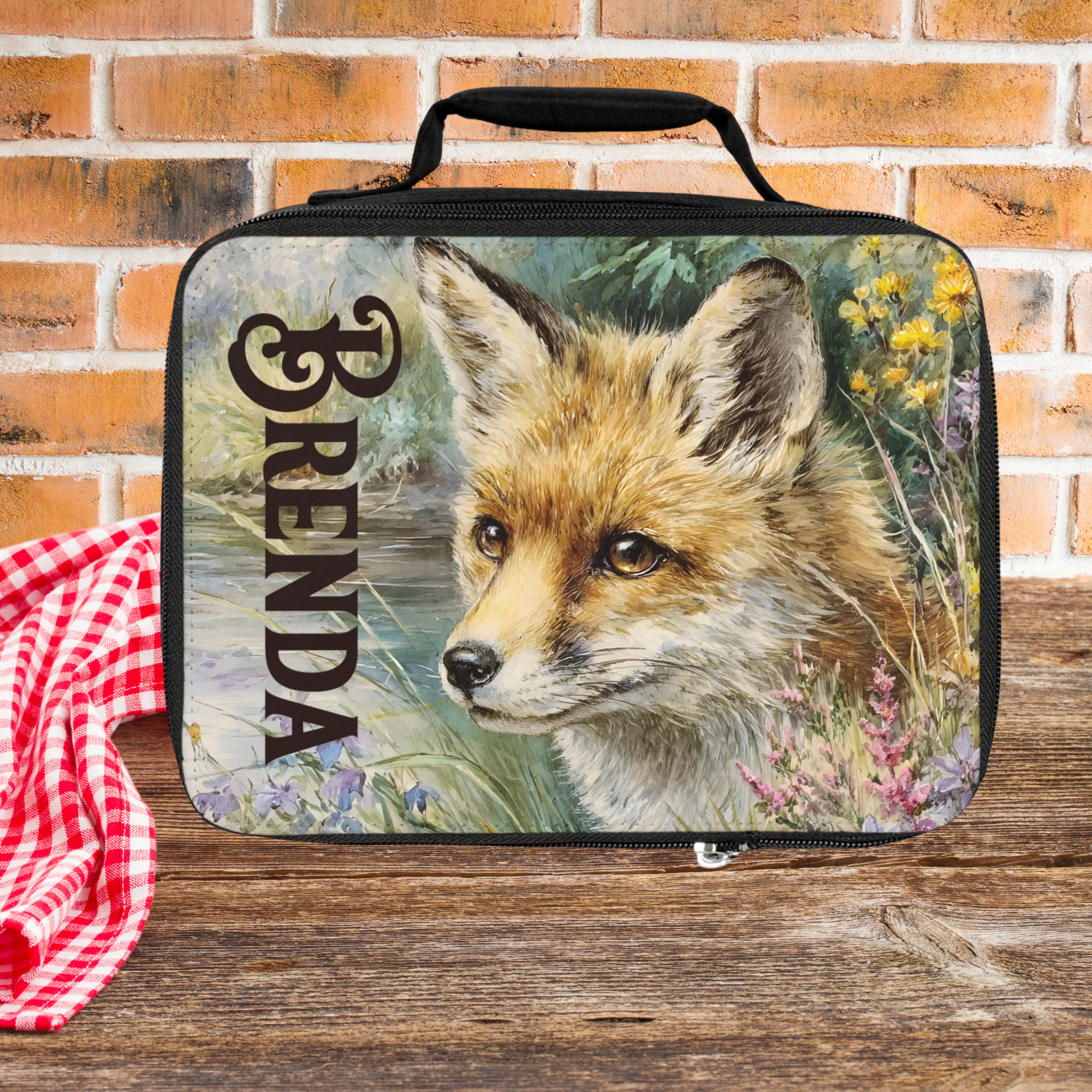 Lunch bag with a fox design and the name 'Brenda' on a wooden picnic table against a brick wall background