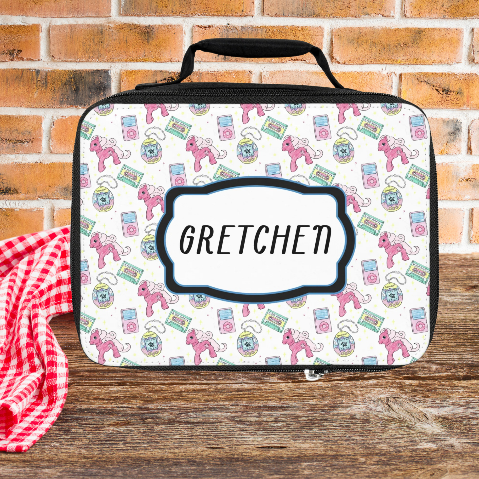 Lunch bag with colorful pattern and 'Gretchen' custom text on a picnic table against a brick wall background
