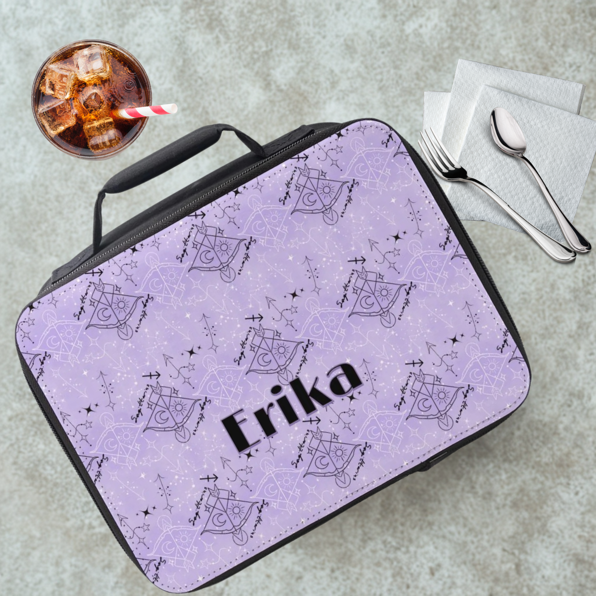 Purple and black lunch bag with 'Erika' printed on it, next to a drink and utensils on a gray table.