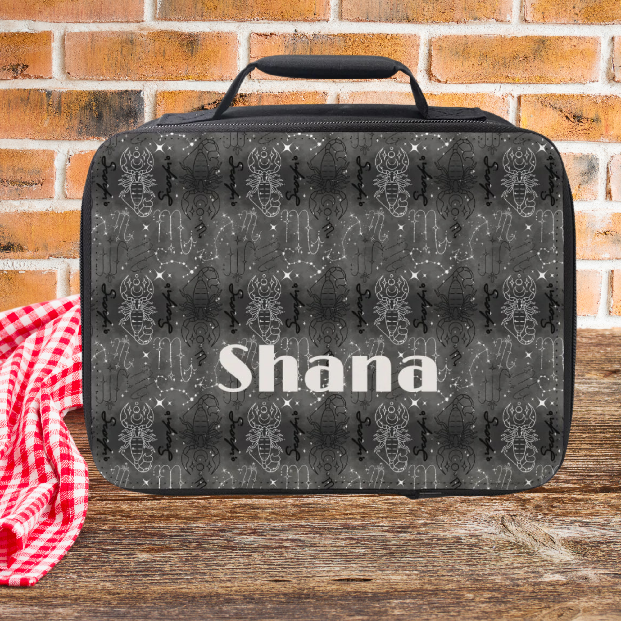 Black Scorpio patterned lunch bag with 'Shana' printed on it, placed on a wooden picnic table with a red checkered cloth and brick wall background.