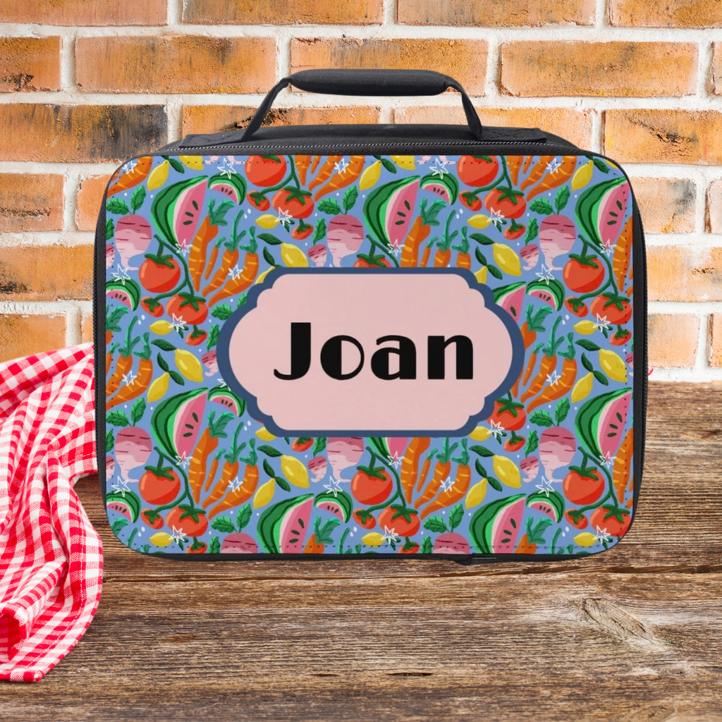Colorful food-patterned lunch bag with 'Joan' custom on a wooden picnic table and brick wall background