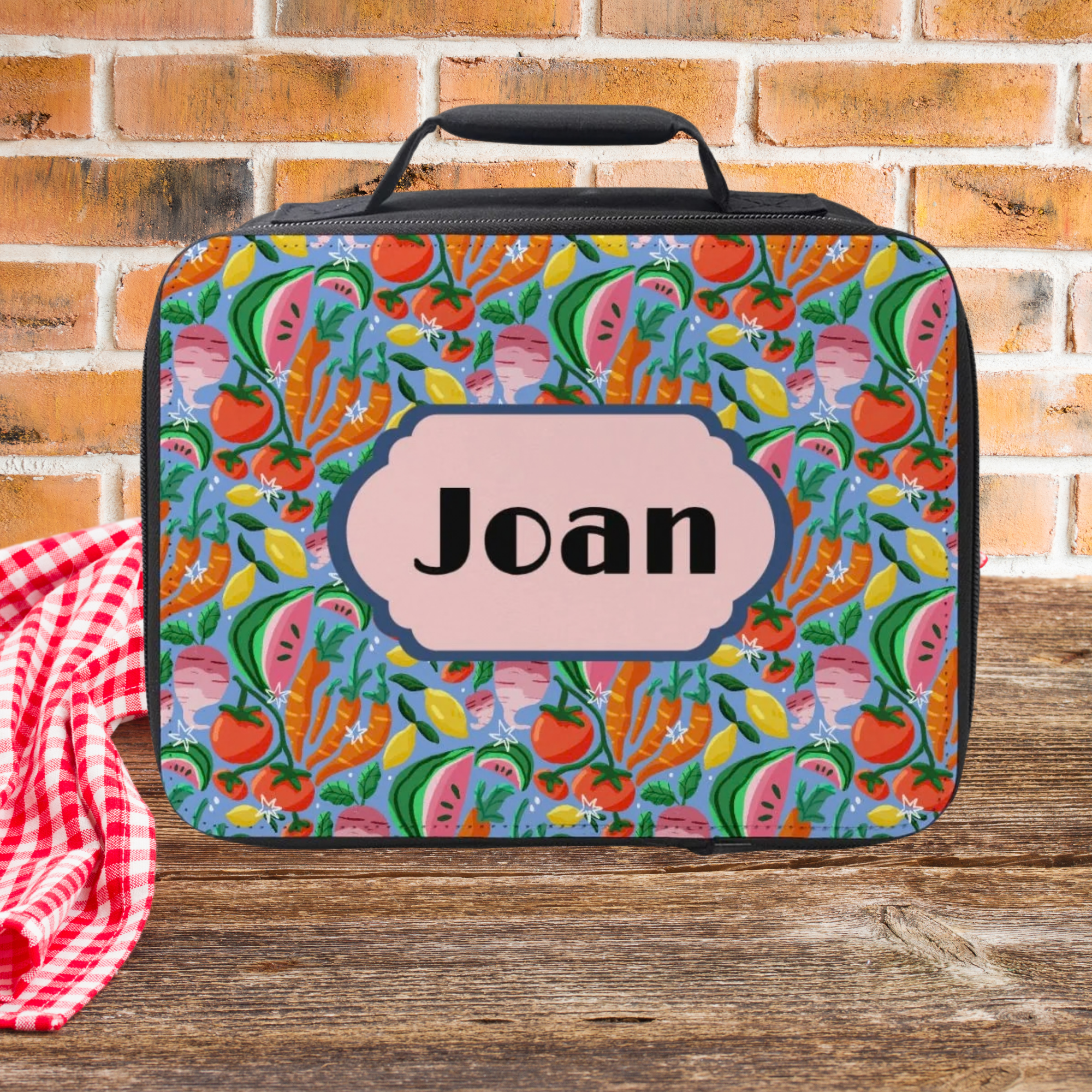Colorful food-patterned lunch bag with 'Joan' custom on a wooden picnic table and brick wall background
